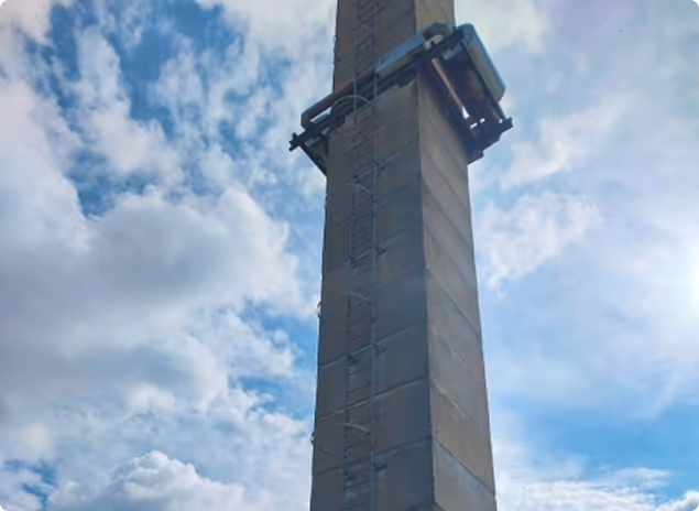 Ein hoher, betonierter Turm mit einer Metallleiter und einer großen Lampe oben, bei bewölktem Himmel.