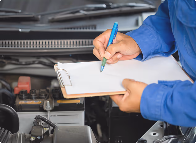 Techniker in blauer Arbeitskleidung macht Notizen auf einem Klemmbrett vor geöffnetem Motorraum eines Fahrzeugs.