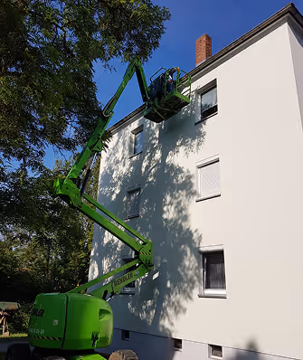 Grüne Arbeitsbühne hebt zwei Personen an eine Fensteröffnung eines weißen dreigeschossigen Gebäudes unter blauem Himmel.