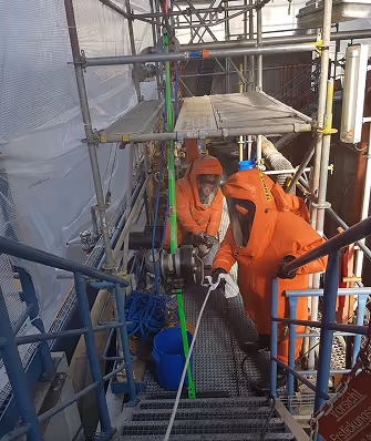 Zwei Arbeiter in orangefarbenen Schutzanzügen mit Atemschutzmaske führen Arbeiten auf einem Gerüst durch.
