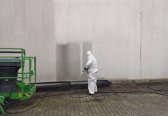 Person im weißen Schutzanzug reinigt mit Hochdruckreiniger eine große, schmutzige Außenwand.