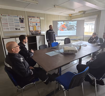 Fünf Männer in dunklen Arbeitsuniformen sitzen und stehen in einem Raum, während einer eine Präsentation mit einem Projektor zeigt.