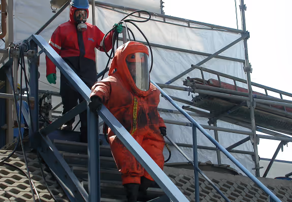 Zwei Personen in Schutzanzügen auf einer Metalltreppe; die vordere Person trägt einen orangefarbenen, gasdichten Schutzanzug mit durchsichtigem Visier, die hintere Person einen roten und schwarzen Schutzanzug mit Atemschutzmaske.