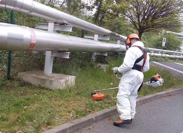 Mitarbeiter in Schutzkleidung und Helm mit Gehörschutz mäht Gras an einer Straßeneinfassung neben großen Rohrleitungen.
