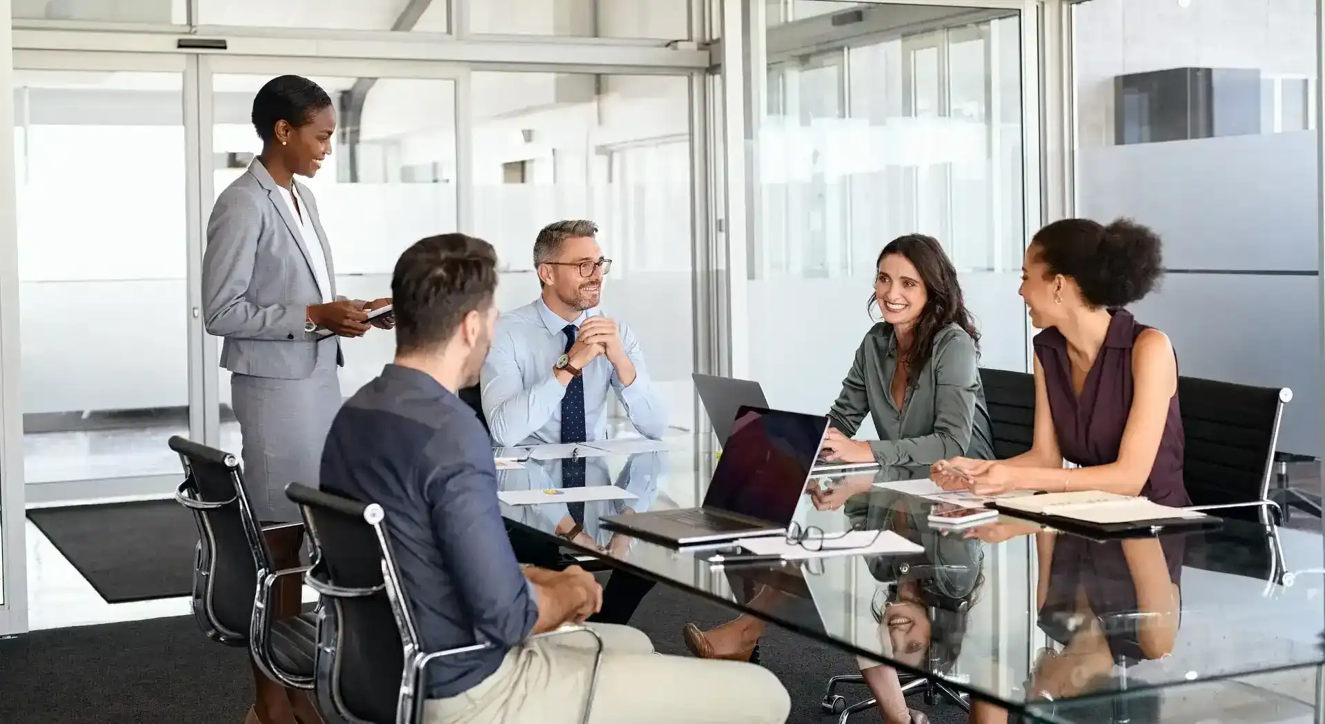 Five diverse business colleagues in a meeting room with a glass table and laptops, smiling and talking.