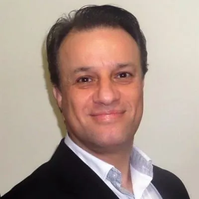 Smiling man with dark hair wearing a black jacket and white shirt against a plain background.