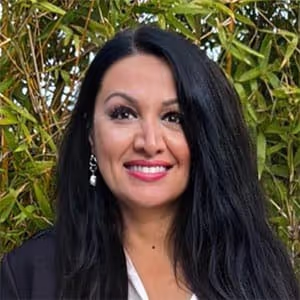 Smiling woman with long black hair, wearing earrings and a dark jacket standing outdoors with green foliage behind.