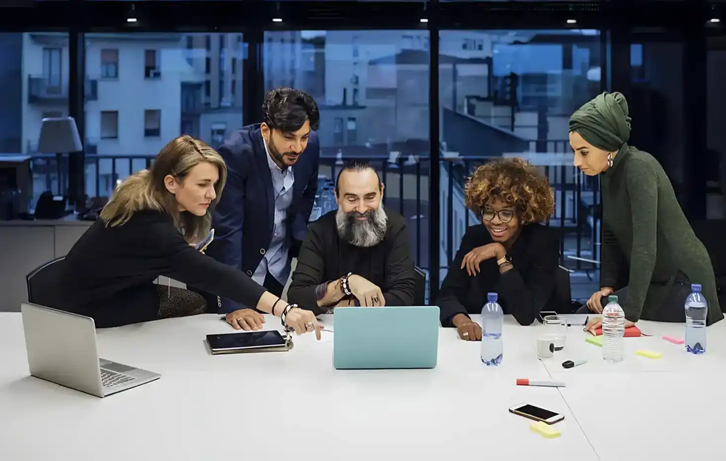 Five diverse coworkers gathered around a table, looking at and discussing a laptop screen.