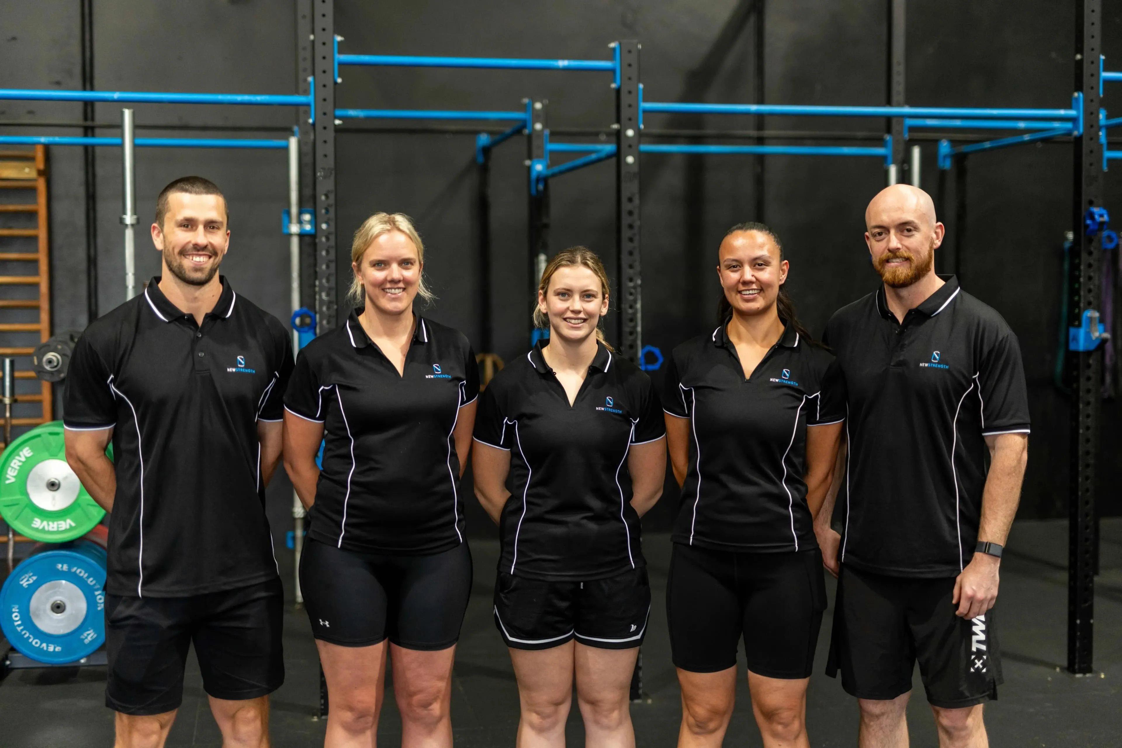 Five fitness trainers standing side by side in a gym wearing matching black polo shirts with a blue and white logo.