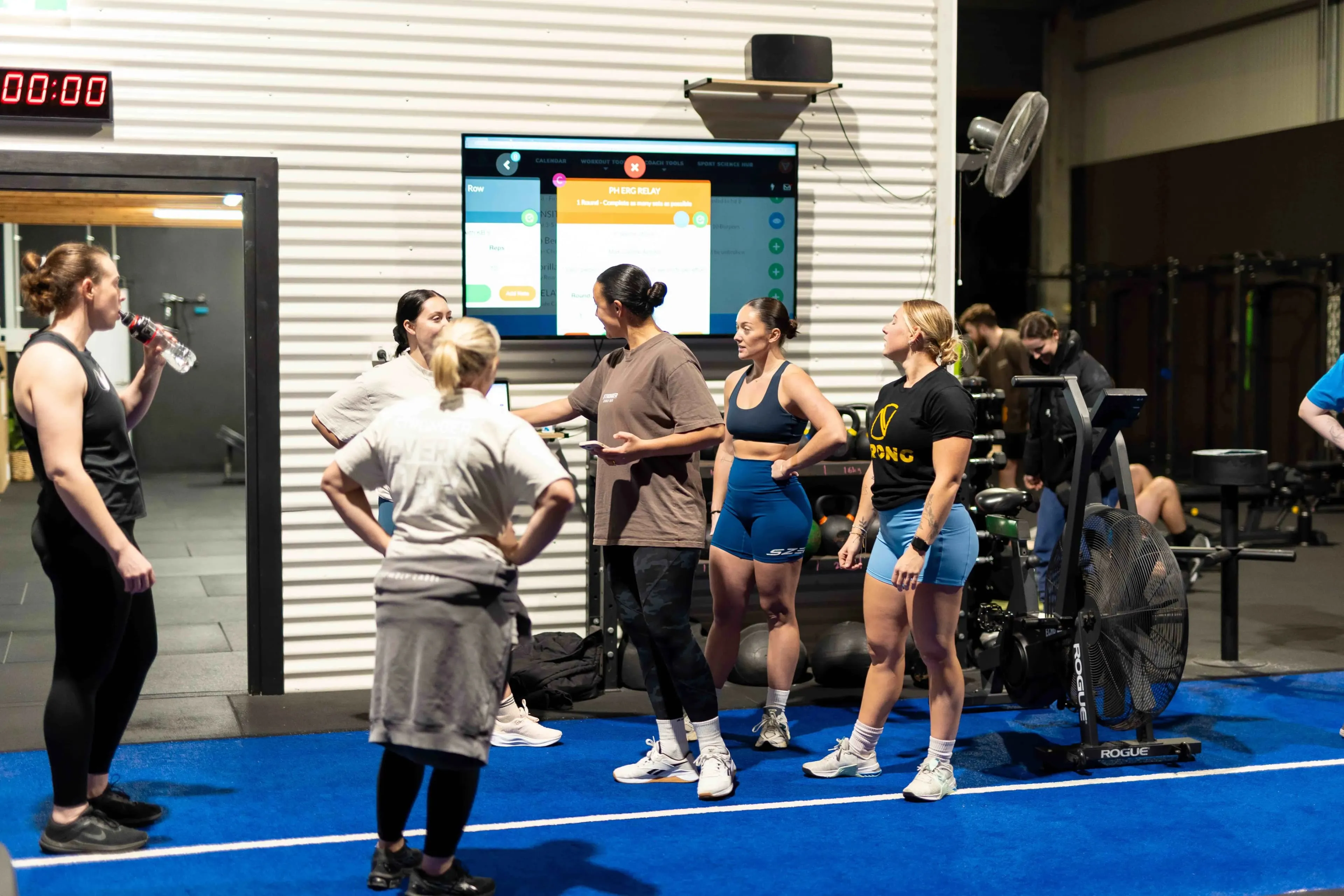 Group of people in athletic wear gathered in a gym, listening to a person explaining a workout displayed on a screen.
