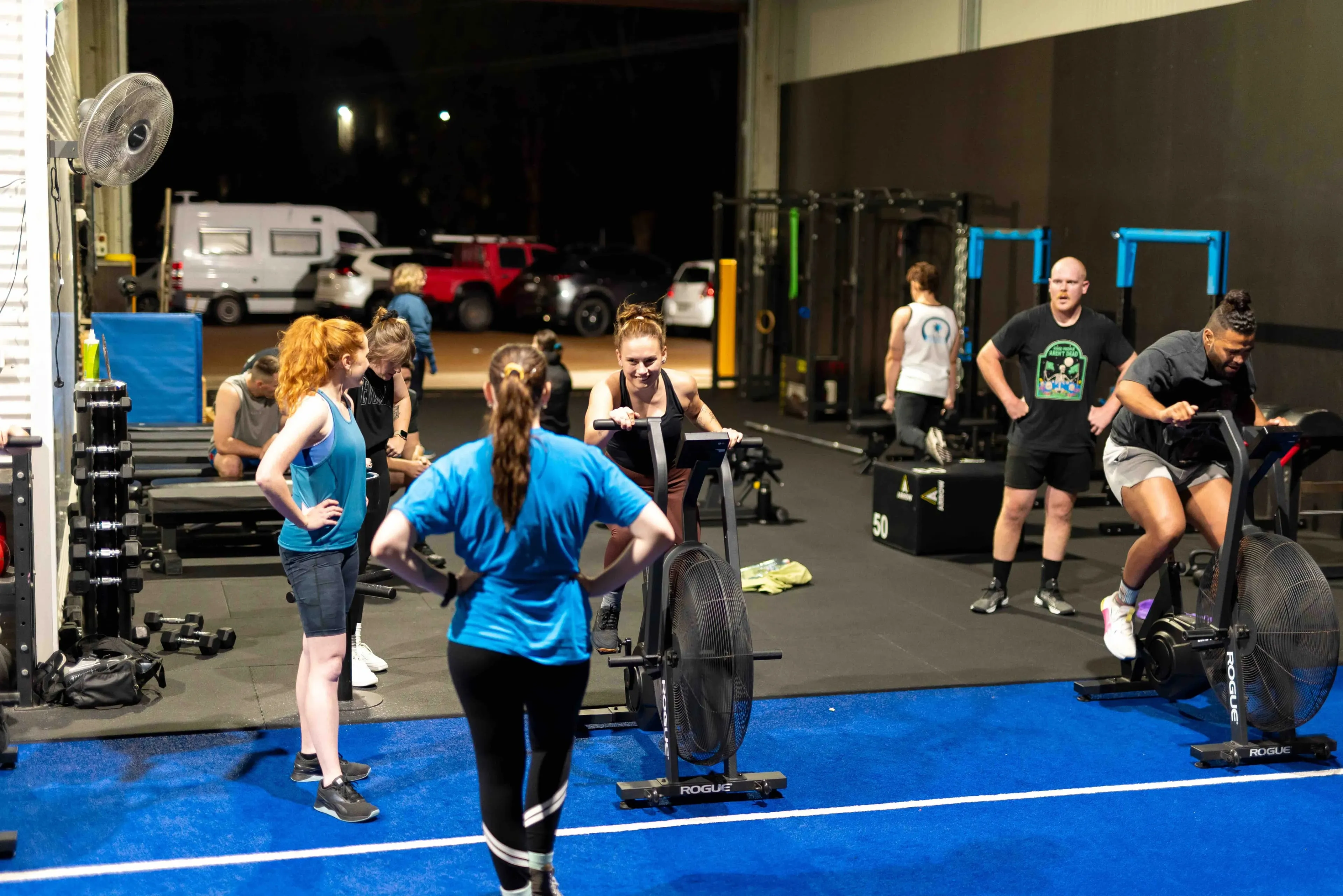 Group of people exercising indoors at night, some using stationary air bikes and others standing or stretching on a blue mat.