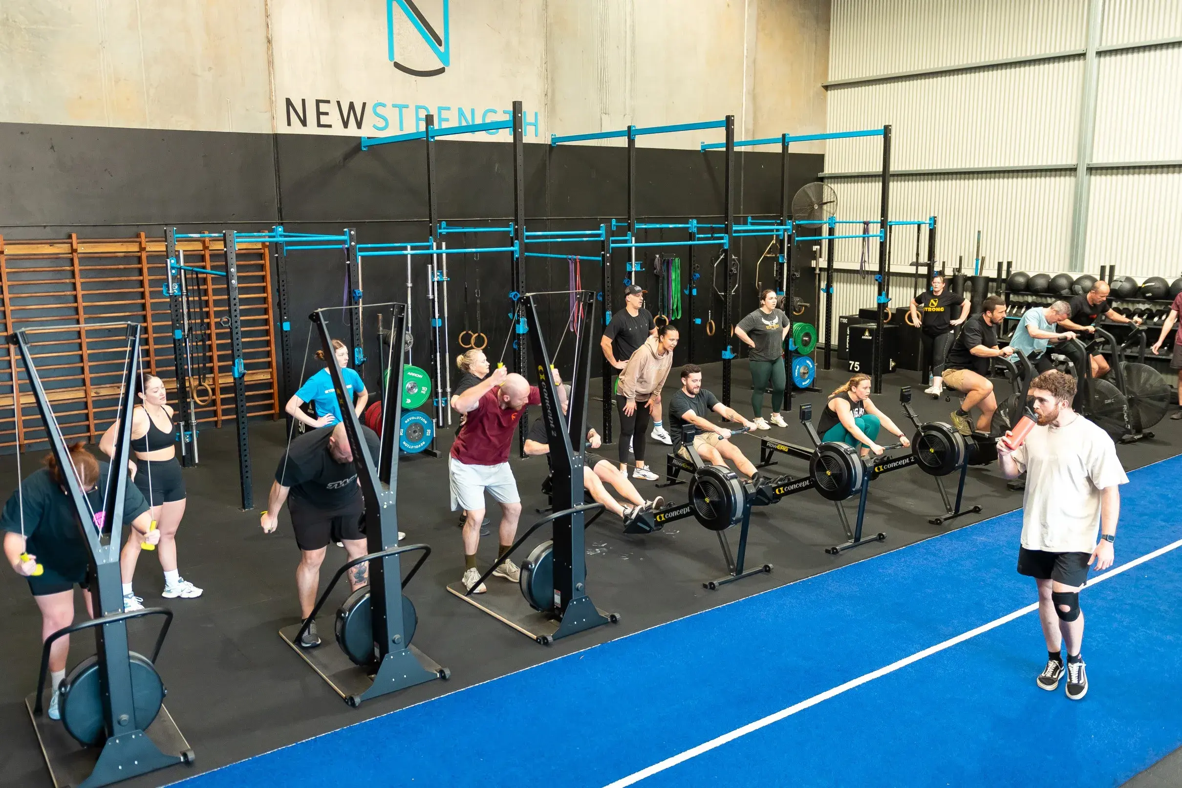 Group of people exercising on rowing machines and resistance cable machines in a gym with blue flooring and black equipment frames.