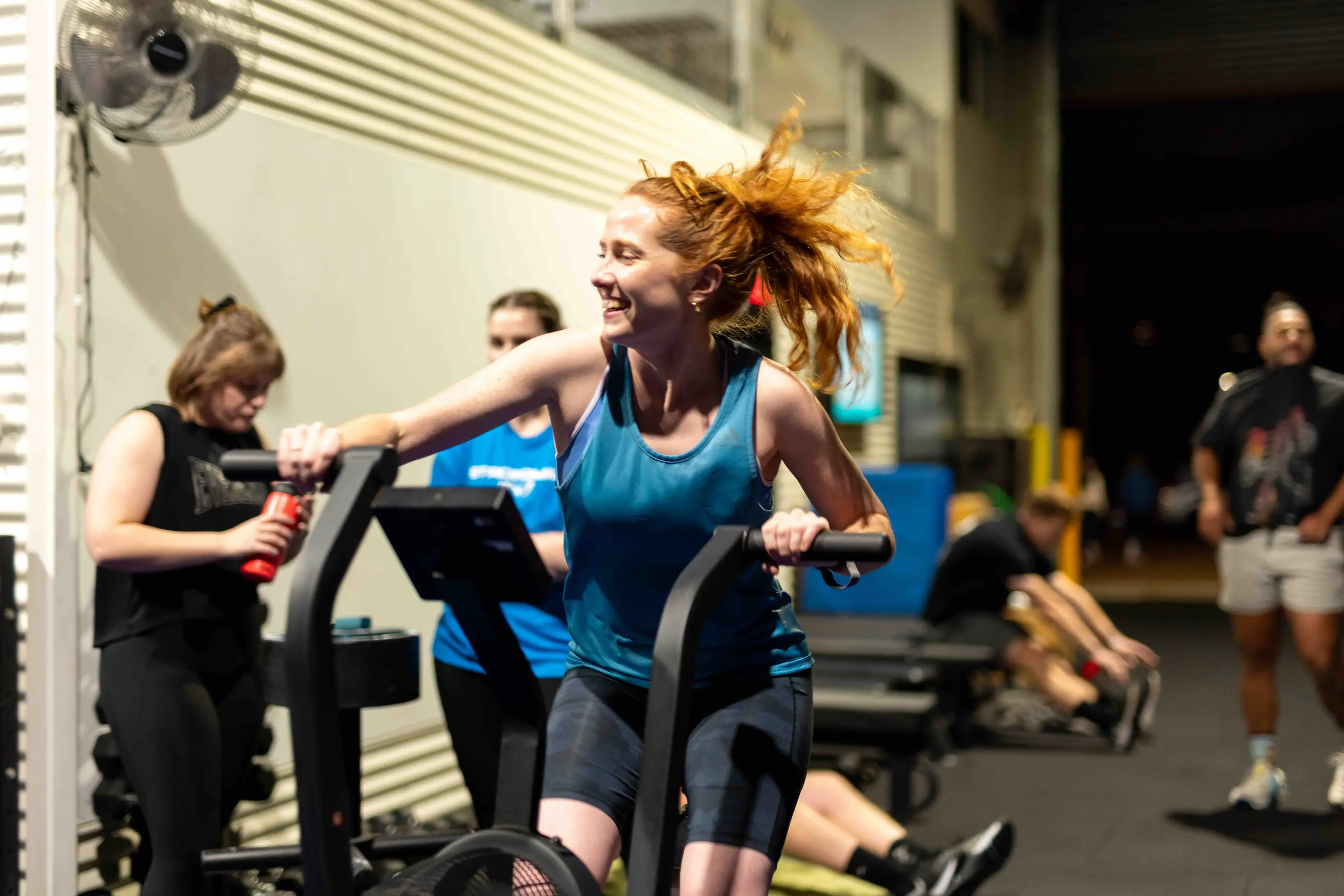 Smiling woman with red hair exercising on a stationary bike in a gym with other people around.