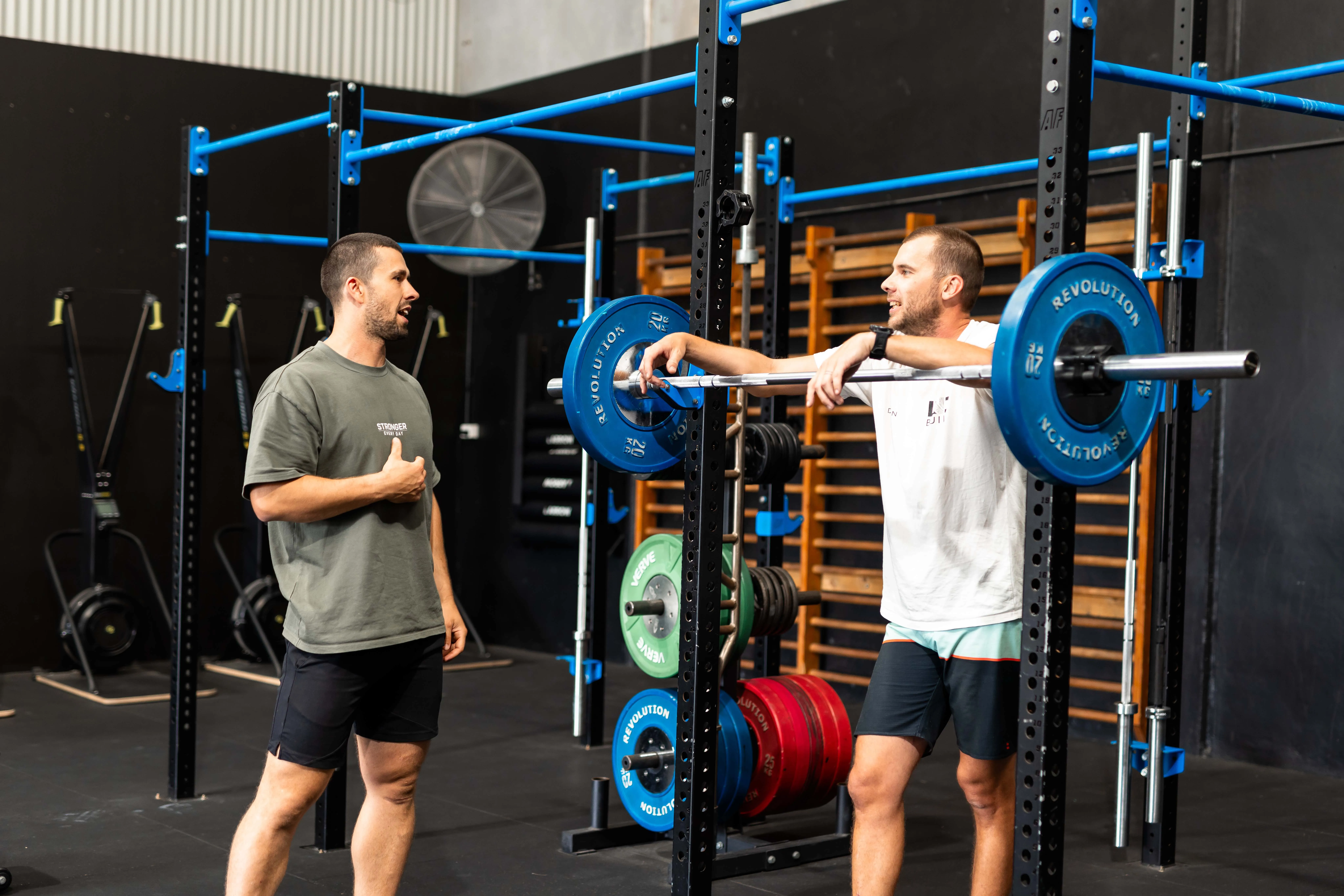 Two men talking in a gym next to a barbell loaded with blue weight plates on a squat rack.