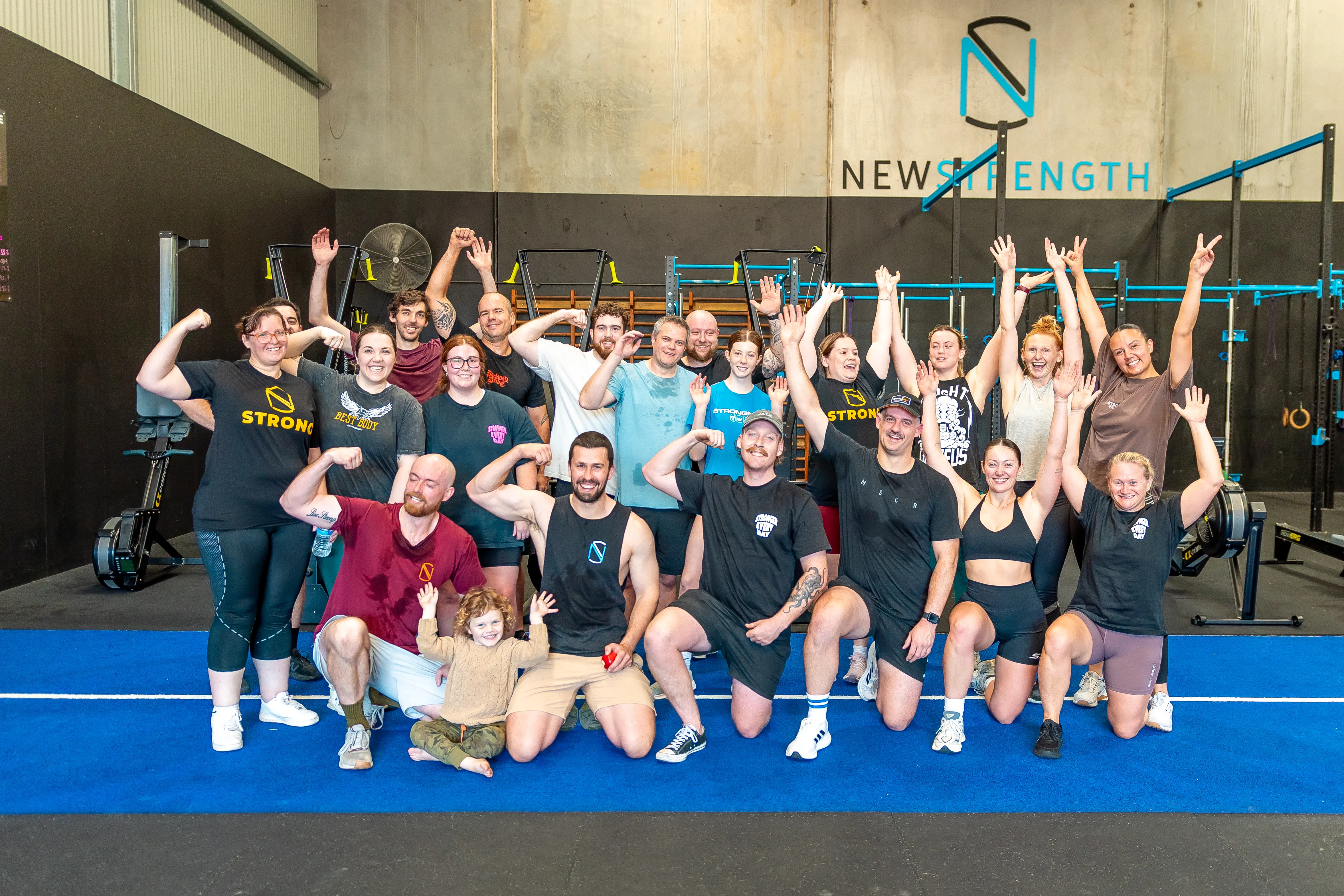 Group of smiling diverse men, women, and a child posing in a gym with workout equipment and a New Strength logo on the wall, some flexing muscles and others raising their arms.