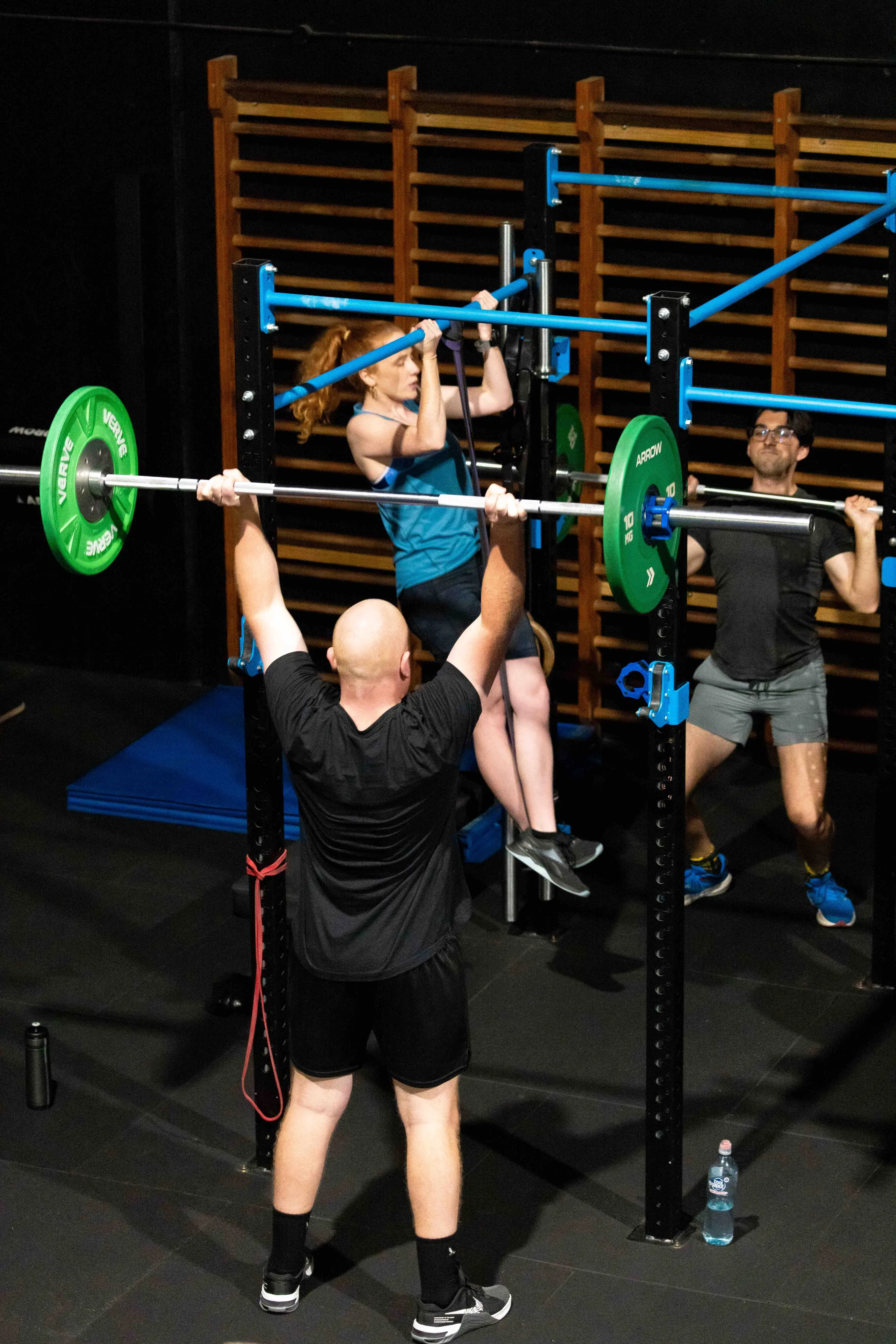 Three people exercising in a gym, one lifting a barbell, another doing a pull-up, and the third lifting a barbell behind the rack.