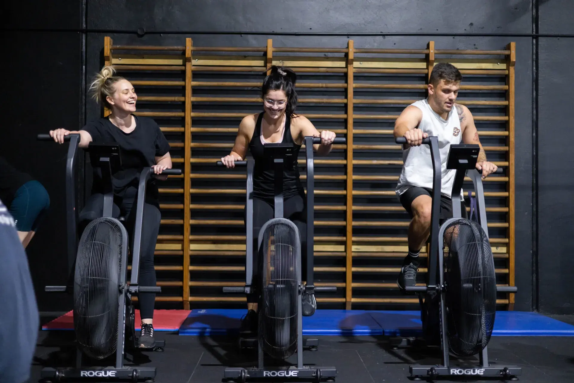 Three people exercising on Rogue stationary air bikes in a gym with wooden wall bars behind them.