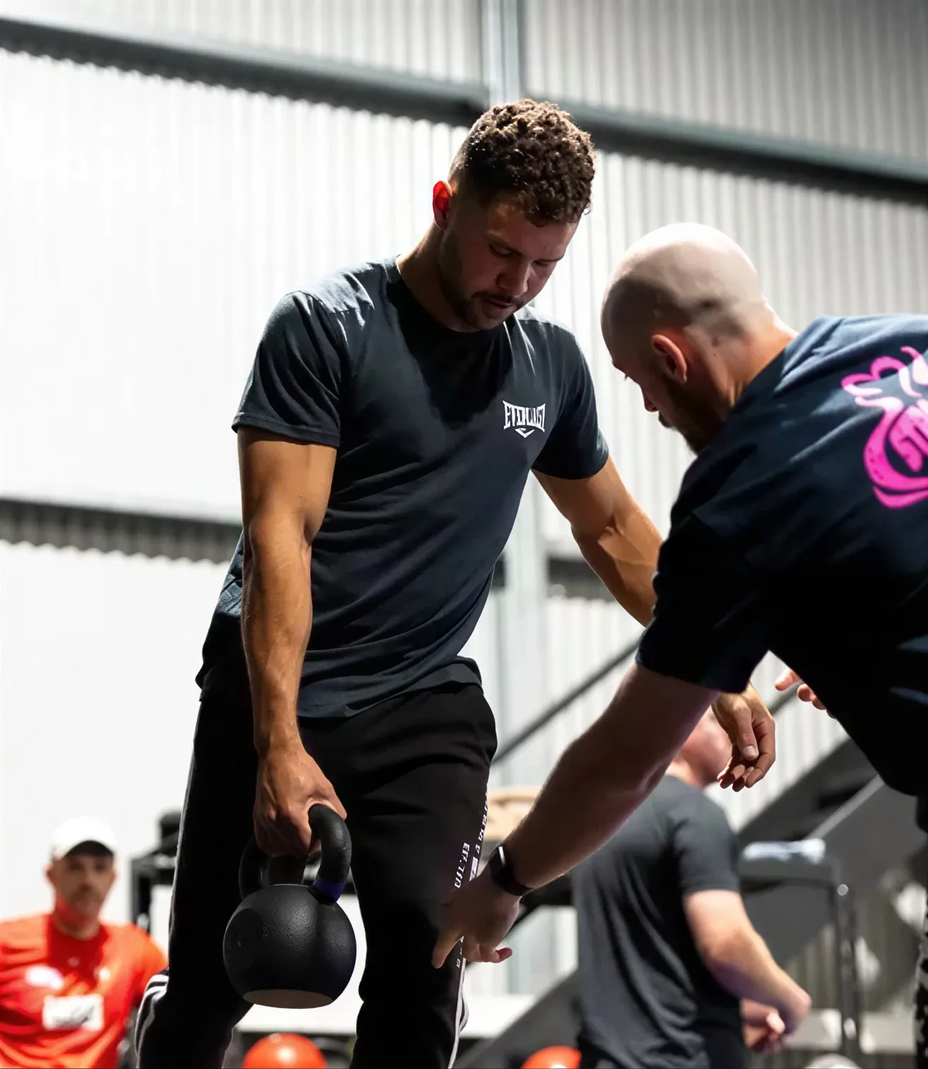 A man lifting a kettlebell receives coaching from another man in a gym setting.