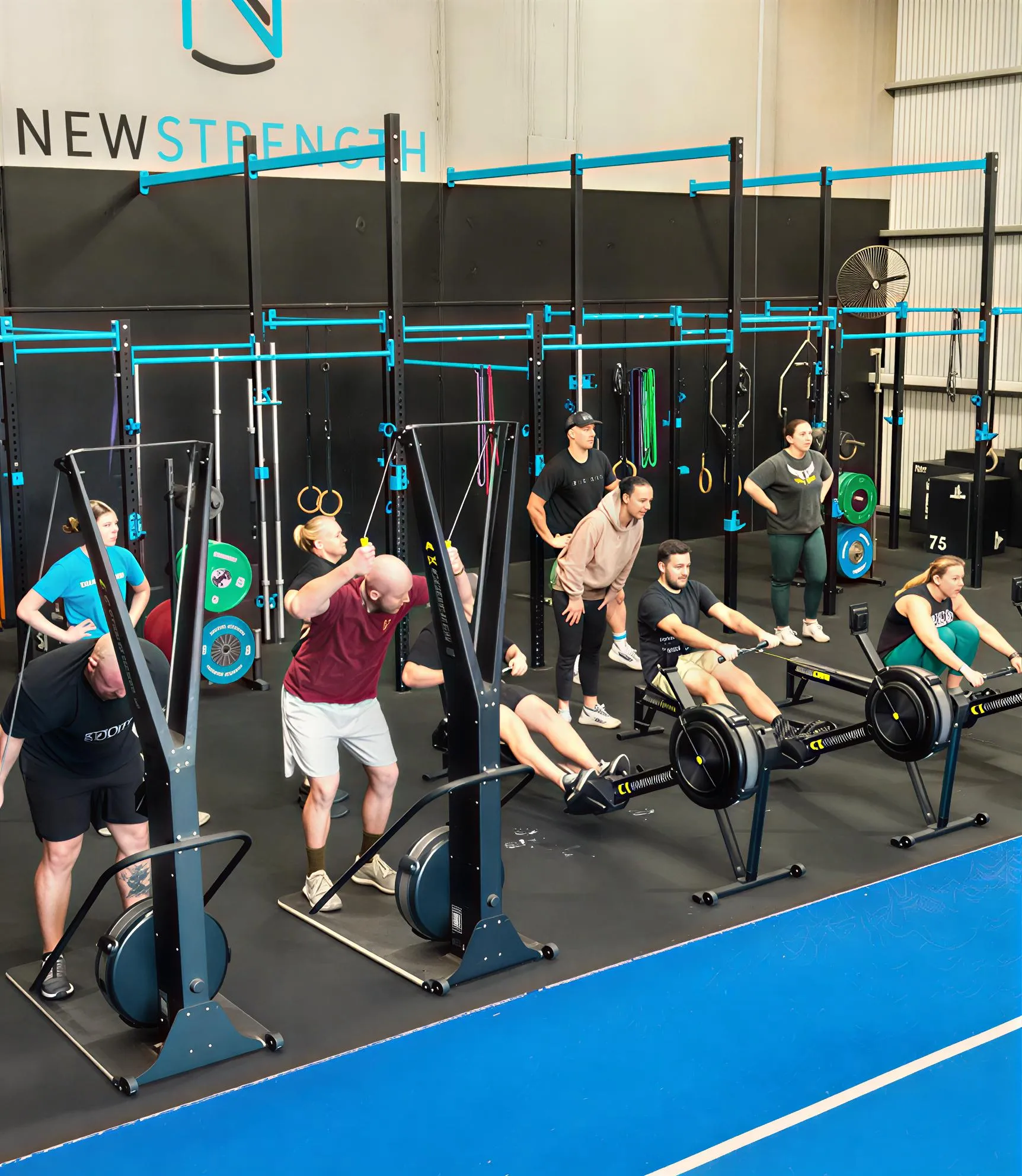 Group of people exercising on rowing machines and air bikes in a gym with New Strength sign on the wall.