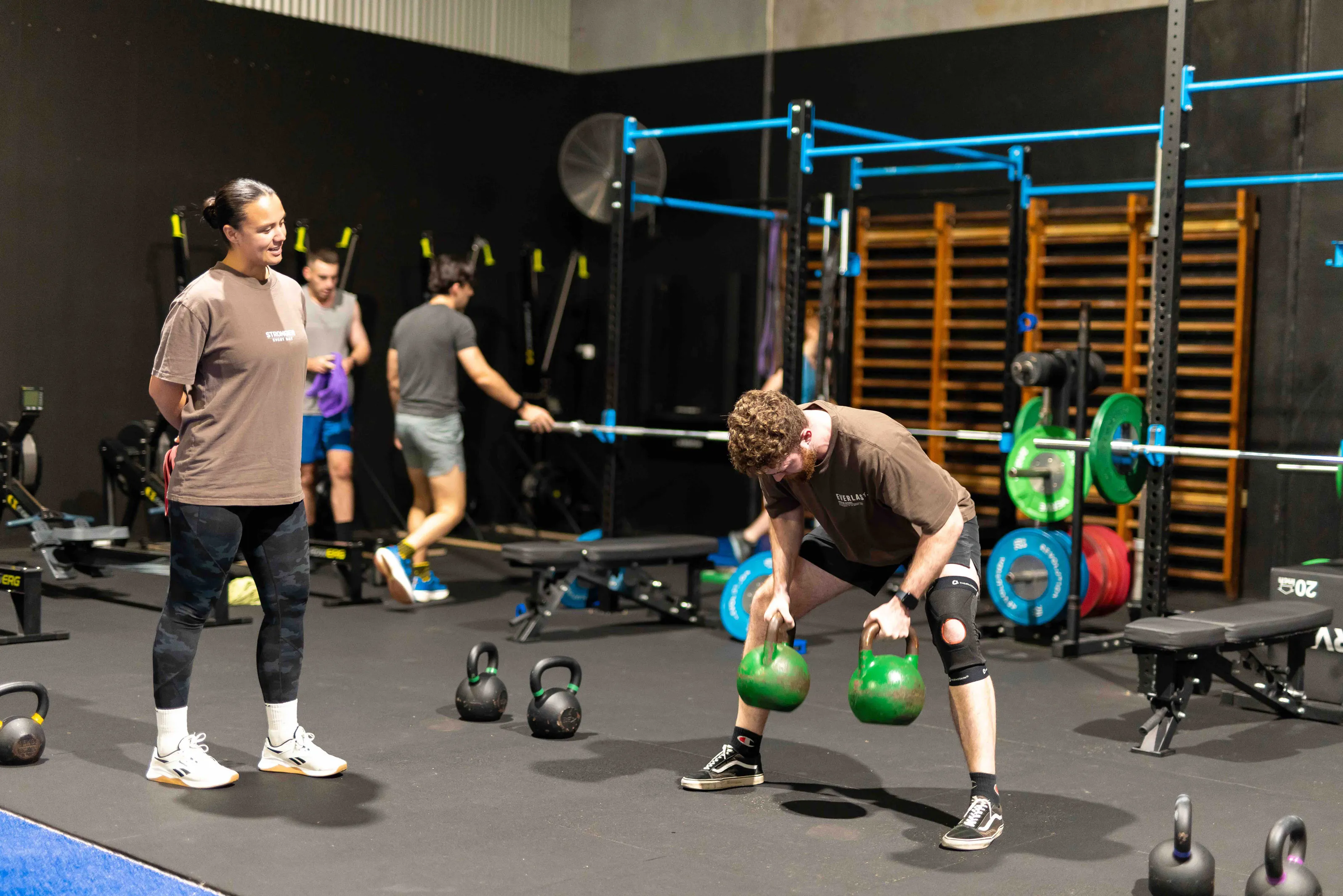Man lifting two green kettlebells in a gym while a woman watches him with a smile.