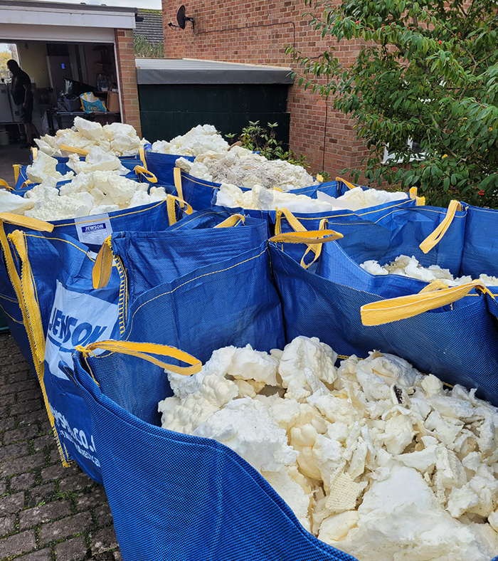 Blue bags filled with large chunks of white sheep wool outside near brick buildings and greenery.