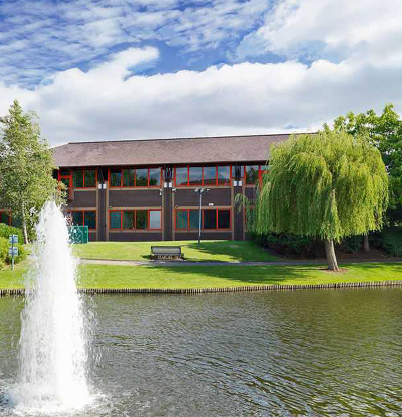 Two-story building with red window frames by a pond with a water fountain and green trees on a sunny day.