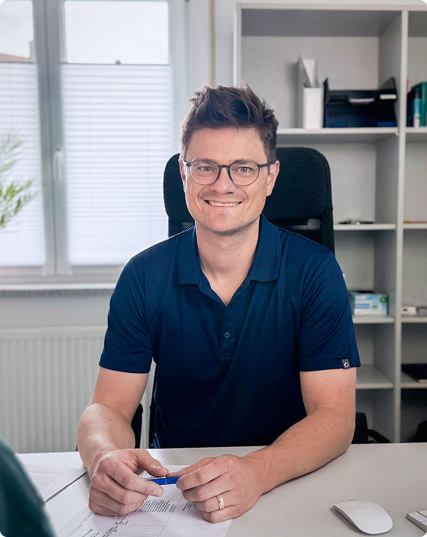 Dr. med. Jonas Elstermann von Elster am Schreibtisch in Büroumgebung mit Regalen und Fenster im Hintergrund.