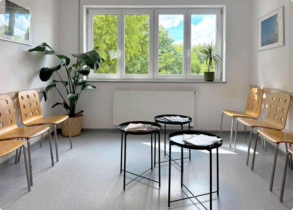 Heller Wartebereich mit Holzstühlen, drei kleinen Tischen, Pflanzen und großem Fenster mit Aussicht auf Bäume.