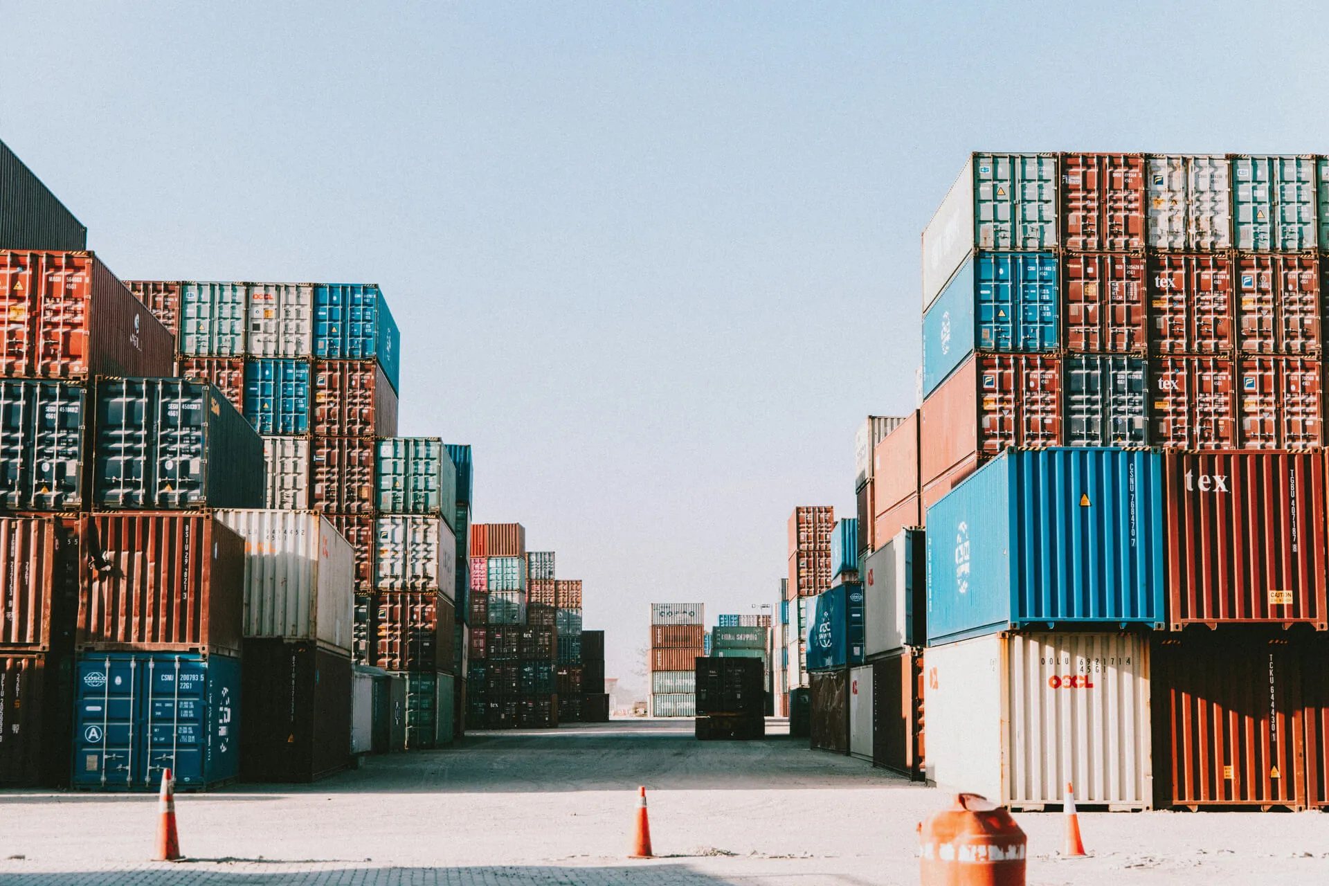 Stacks of colorful shipping containers lined up on both sides of a wide open shipping yard with orange traffic cones in the foreground.