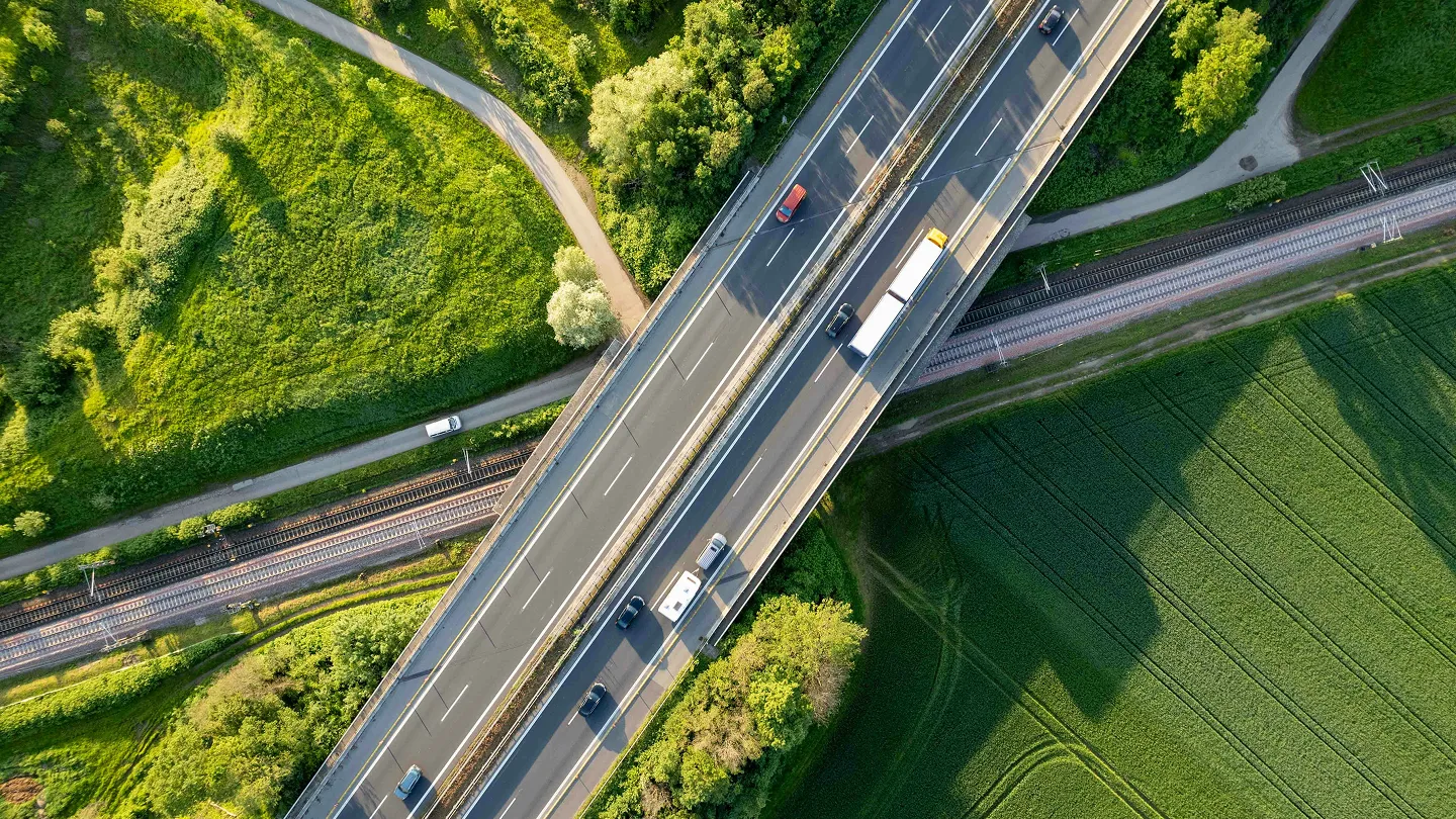 Aerial view of a highway bridge crossing over train tracks, surrounded by green fields and trees.