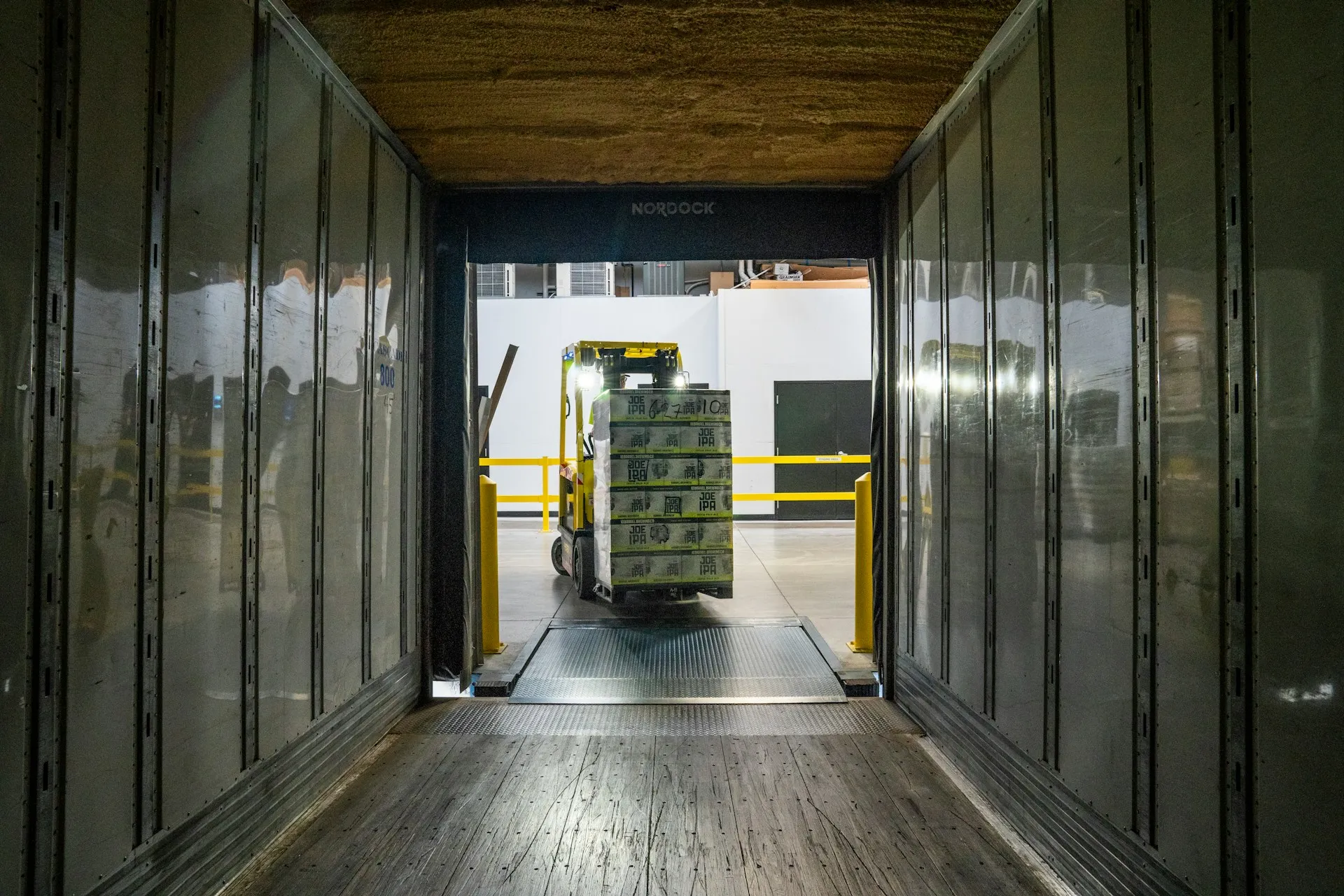 Forklift carrying stacked boxes labeled Joe IPA into a loading dock from inside a truck trailer.