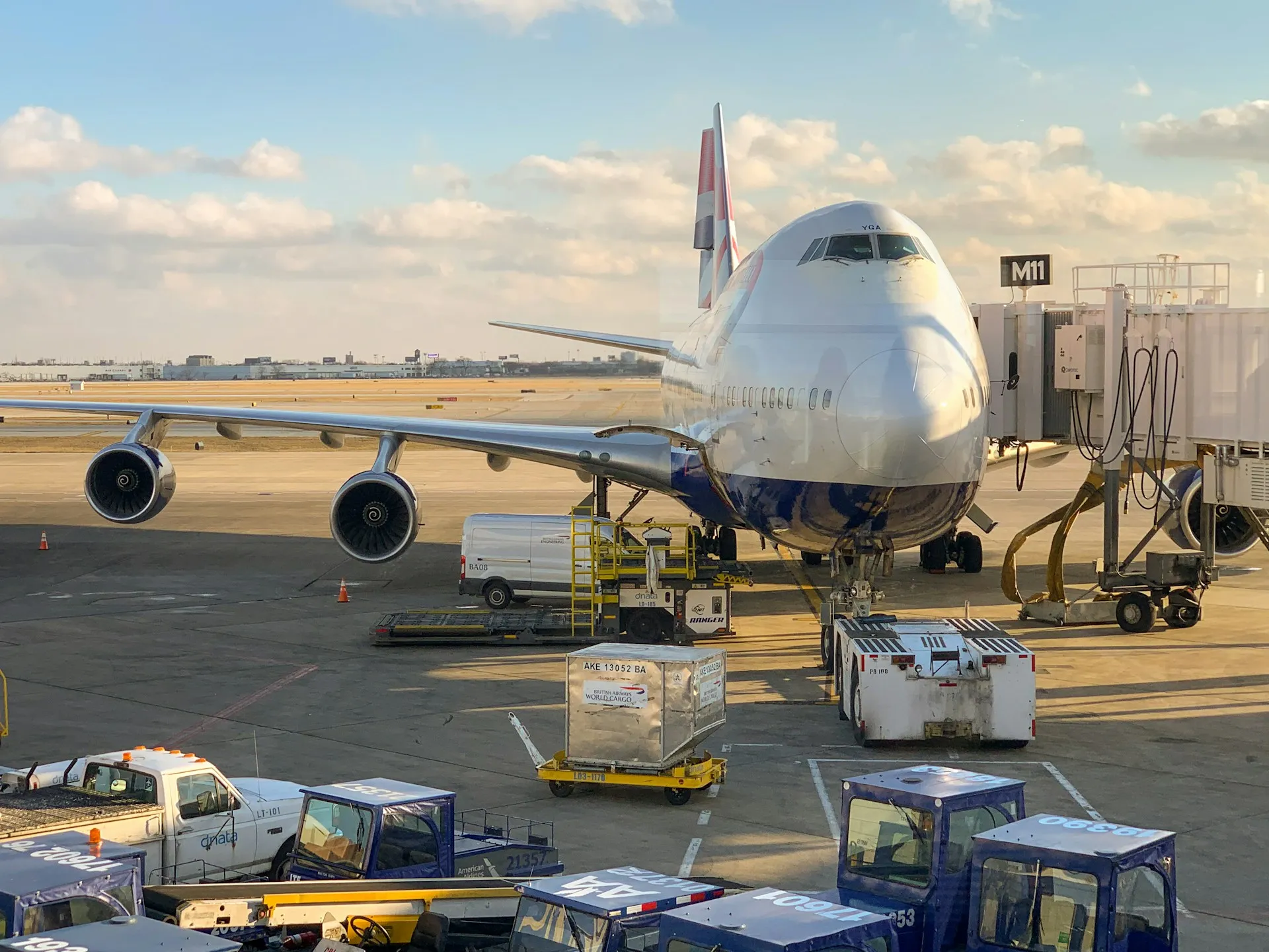 Large commercial airplane docked at airport gate M11 with ground service vehicles and cargo containers nearby.