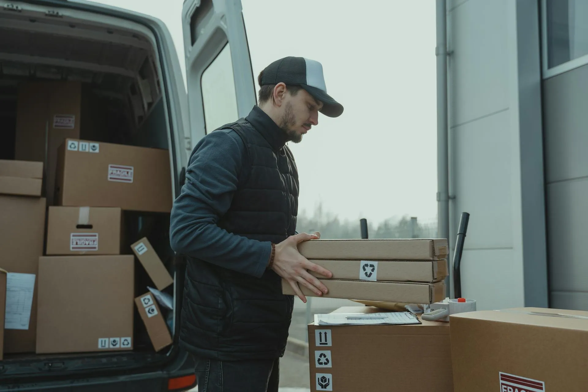 Man wearing a cap and vest unloading flat cardboard boxes from a van and stacking them on a larger box outside a building.