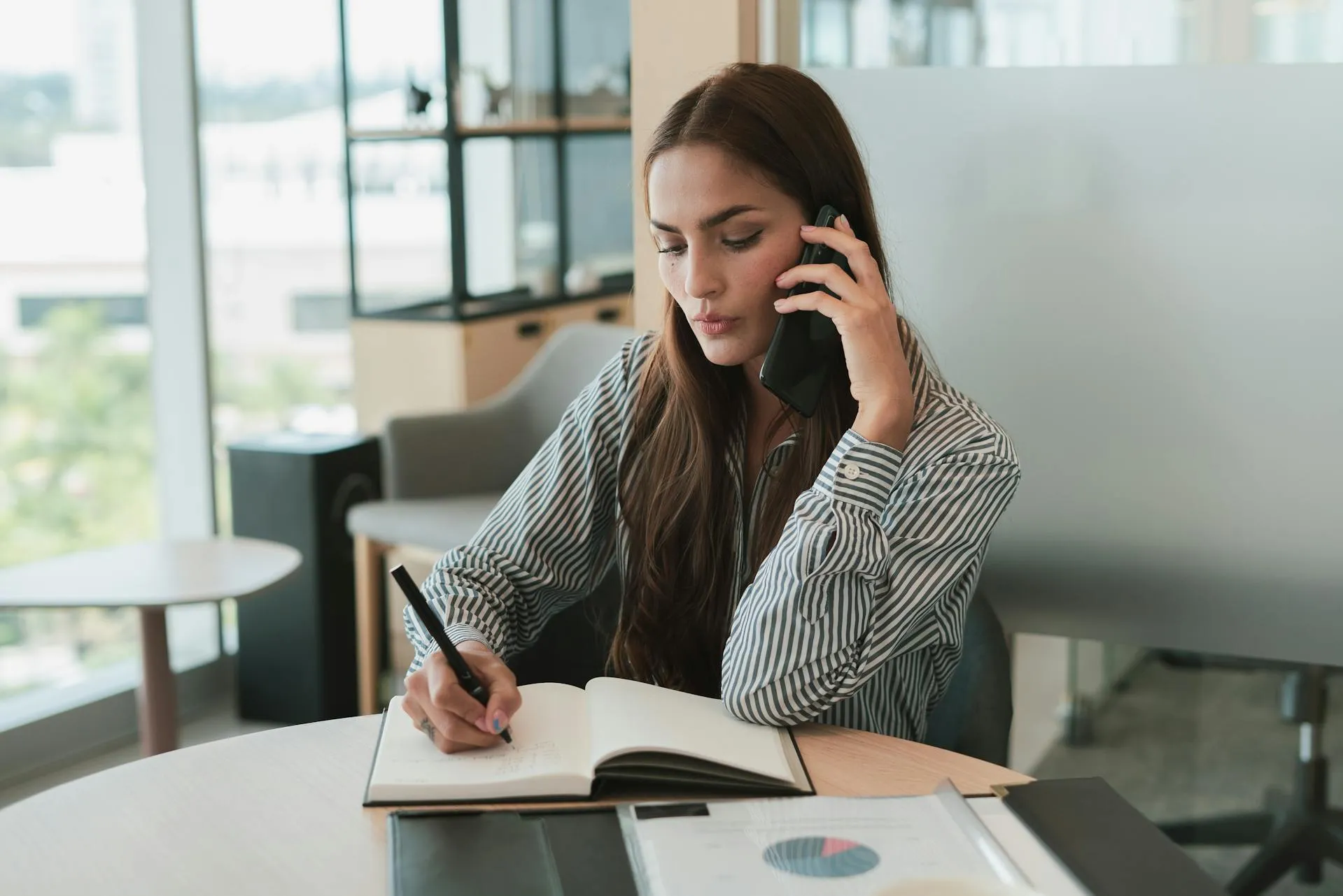 A woman in a striped shirt talks on a smartphone while writing in a notebook at a desk in a modern office.