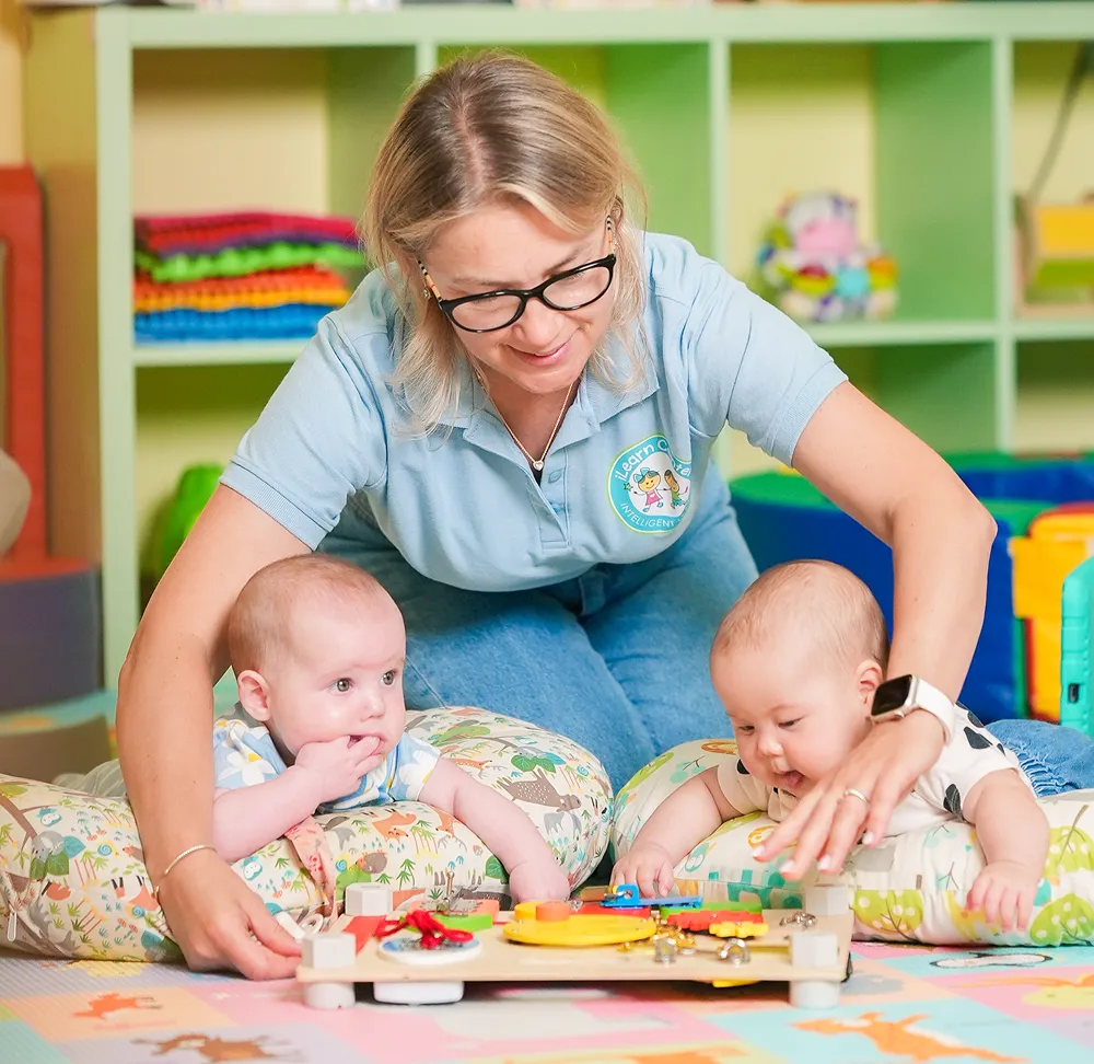 Woman with glasses helps two babies lying on cushions play with a wooden activity board.
