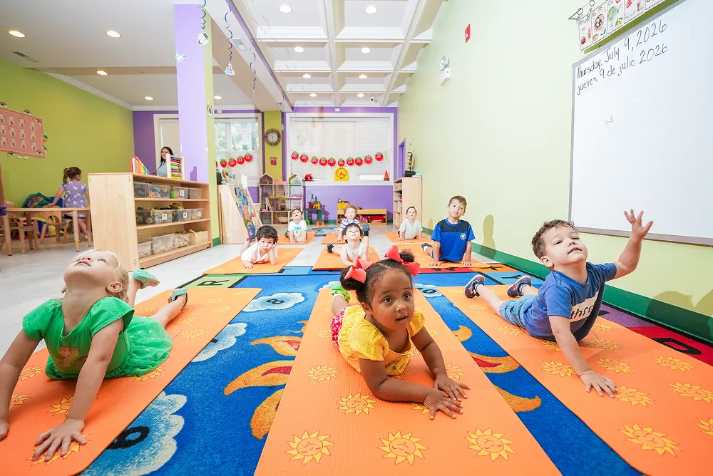 Young children in a classroom doing yoga poses on orange mats on a colorful carpet.