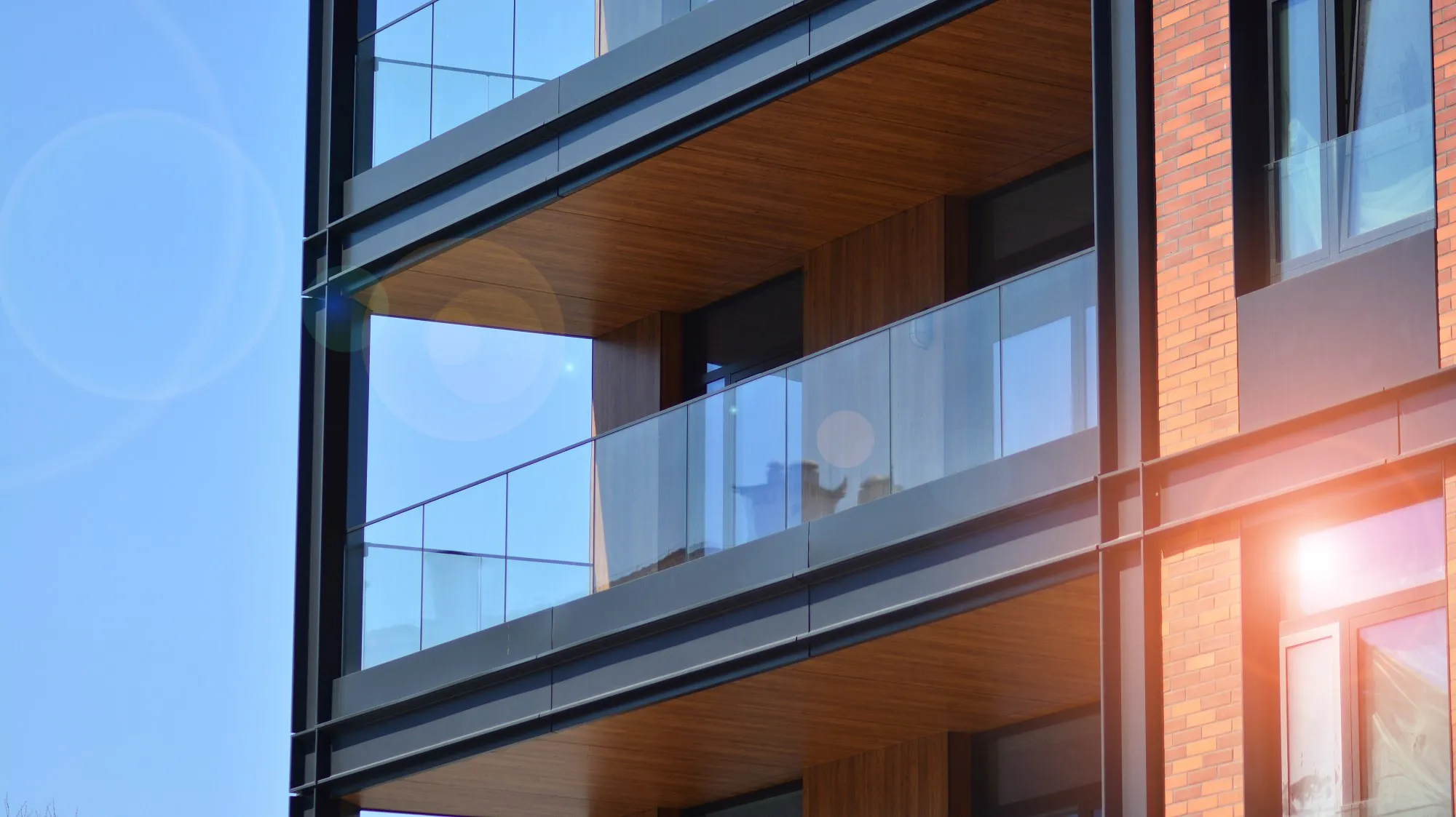 Modern building balcony with glass railing and wood ceiling panels under clear blue sky with sunlight flare.