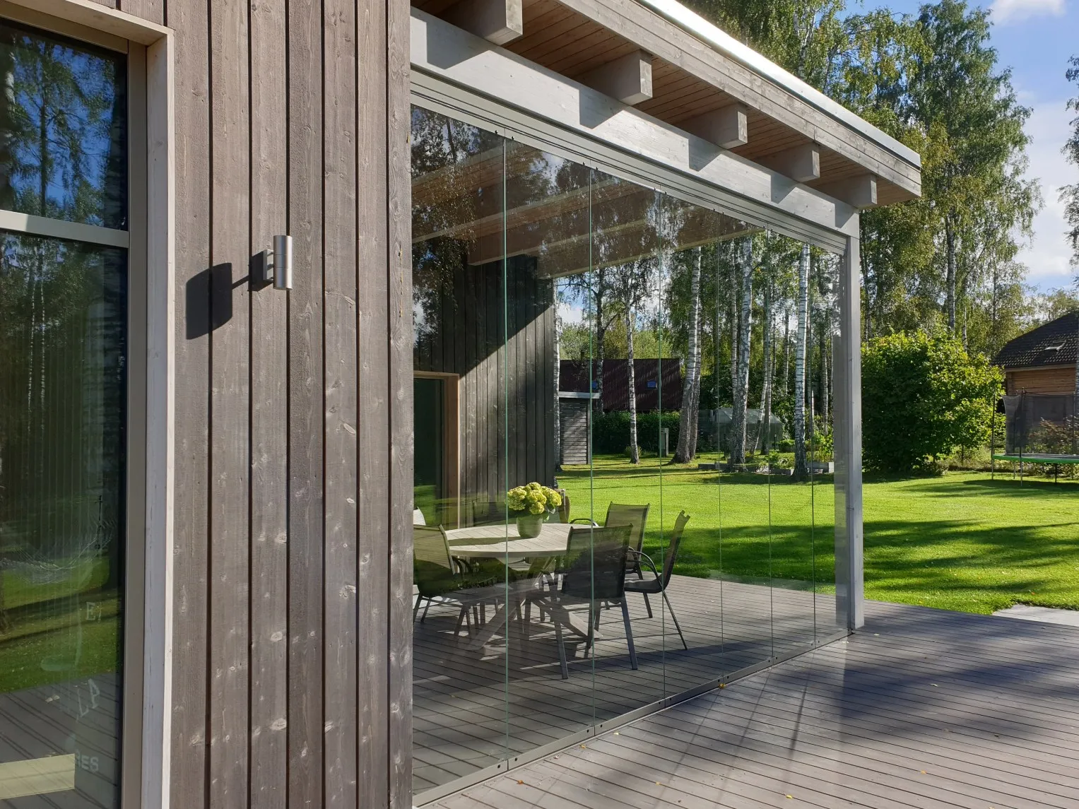 Modern wooden house patio with glass enclosure, outdoor dining table and chairs reflected against green lawn and trees.