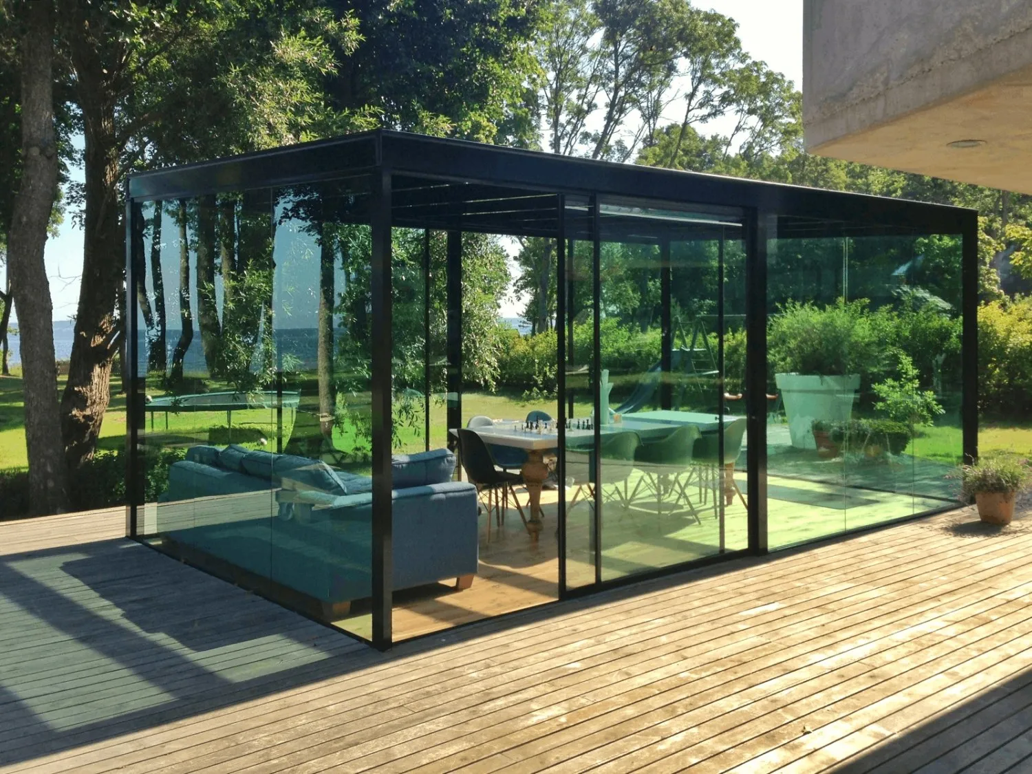Modern glass enclosure on a wooden deck containing a blue sofa, a table with a chessboard, and chairs surrounded by green trees and lawn.