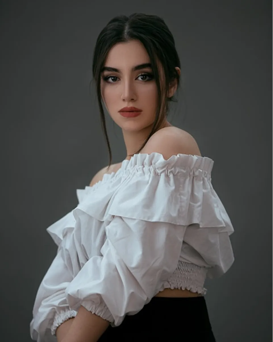 Portrait of a woman with dark hair wearing a white off-the-shoulder ruffled blouse against a gray background.