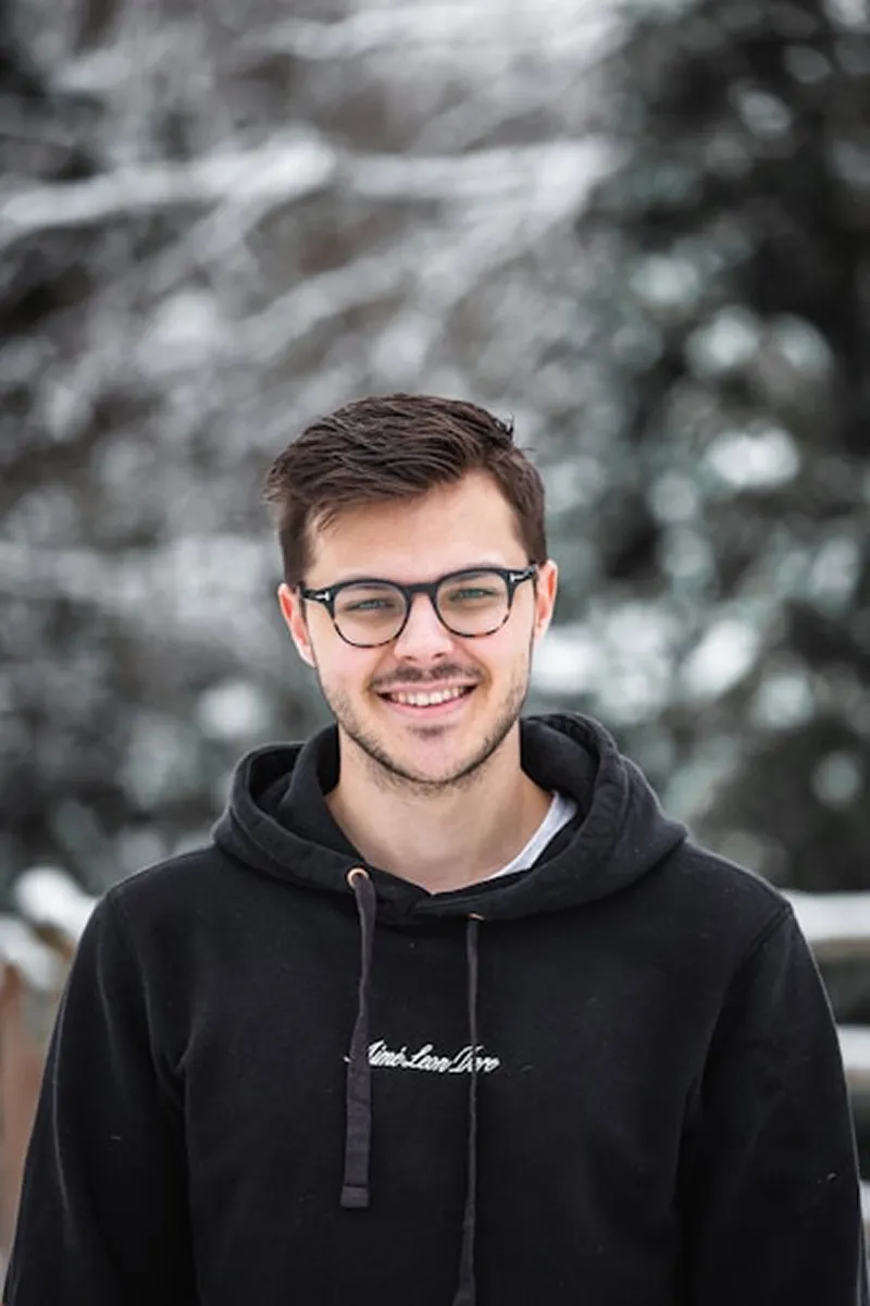 Young man wearing glasses and a black hoodie smiling outdoors with blurred snowy trees in the background.