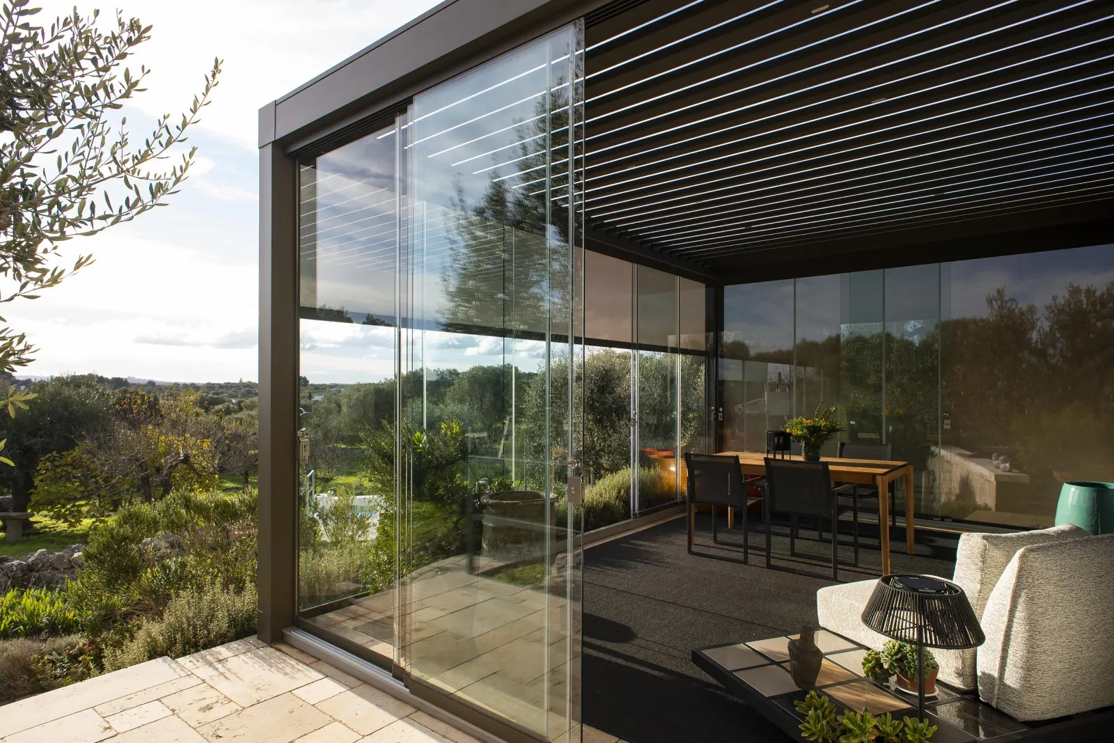 Modern outdoor patio with glass walls, a wooden dining table with chairs, and a seating area overlooking a green garden.