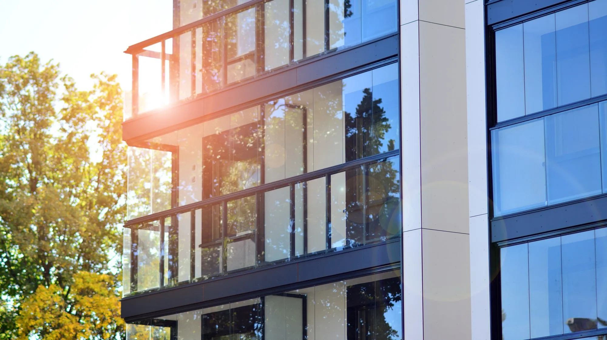 Modern building facade with glass balconies reflecting sunlight and surrounding trees.