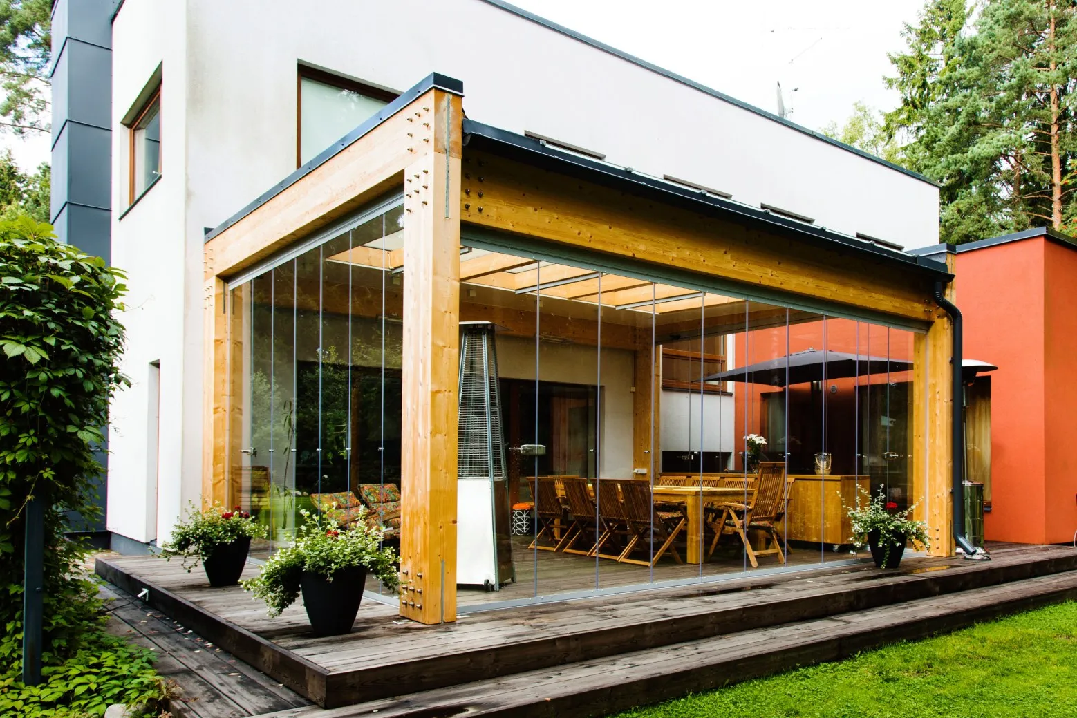 Modern patio with wooden frame and glass walls featuring a dining table and chairs, surrounded by greenery.