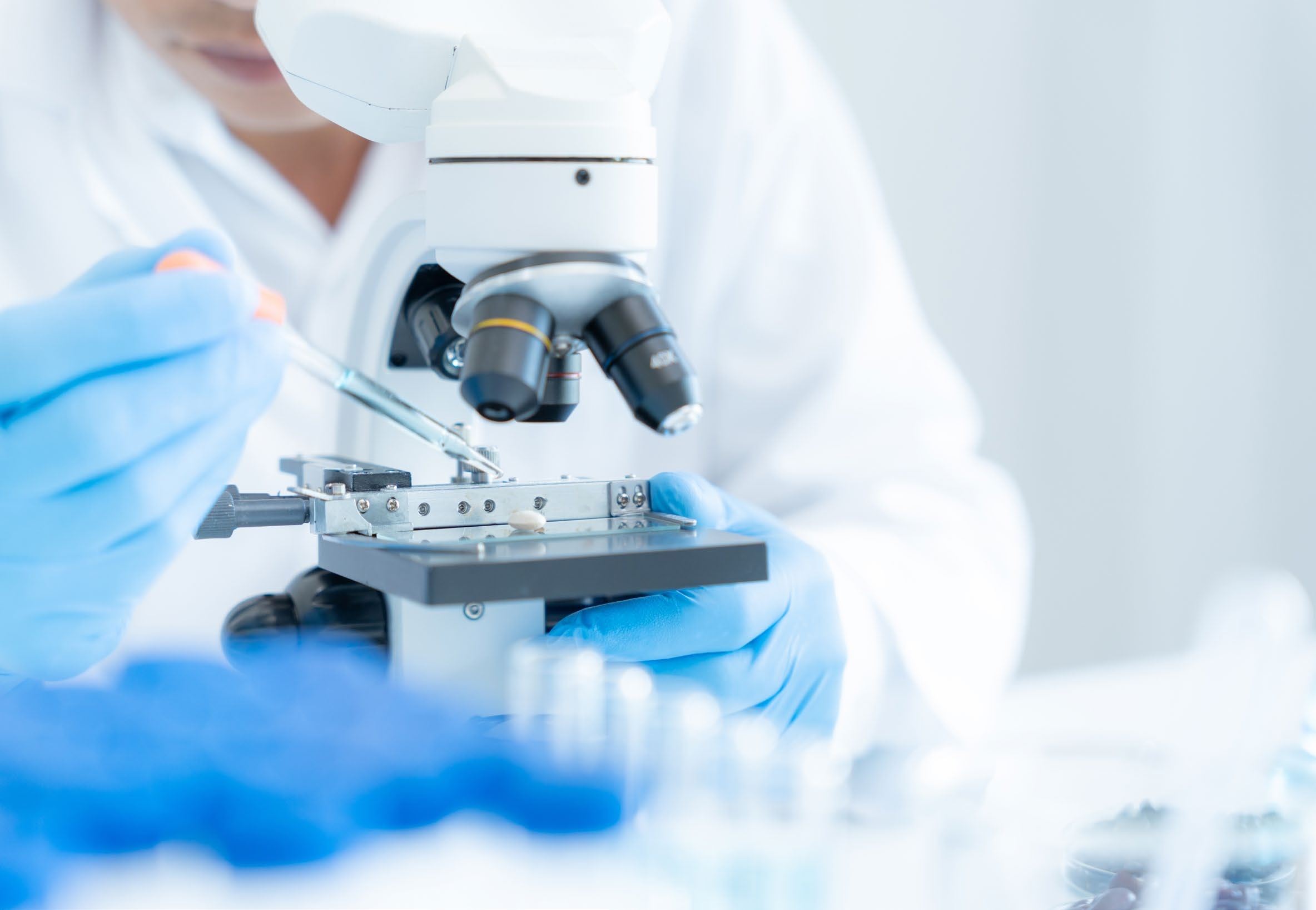 Researcher in lab coat using a pipette on a microscope stage with blurred blue gloves in foreground
