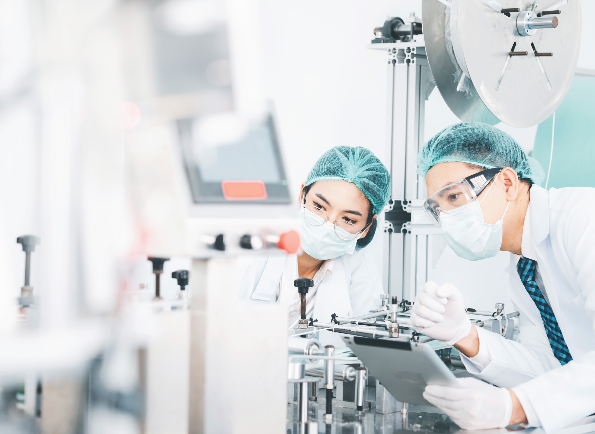 Two researchers in masks and hairnets examining manufacturing equipment in a sterile production facility