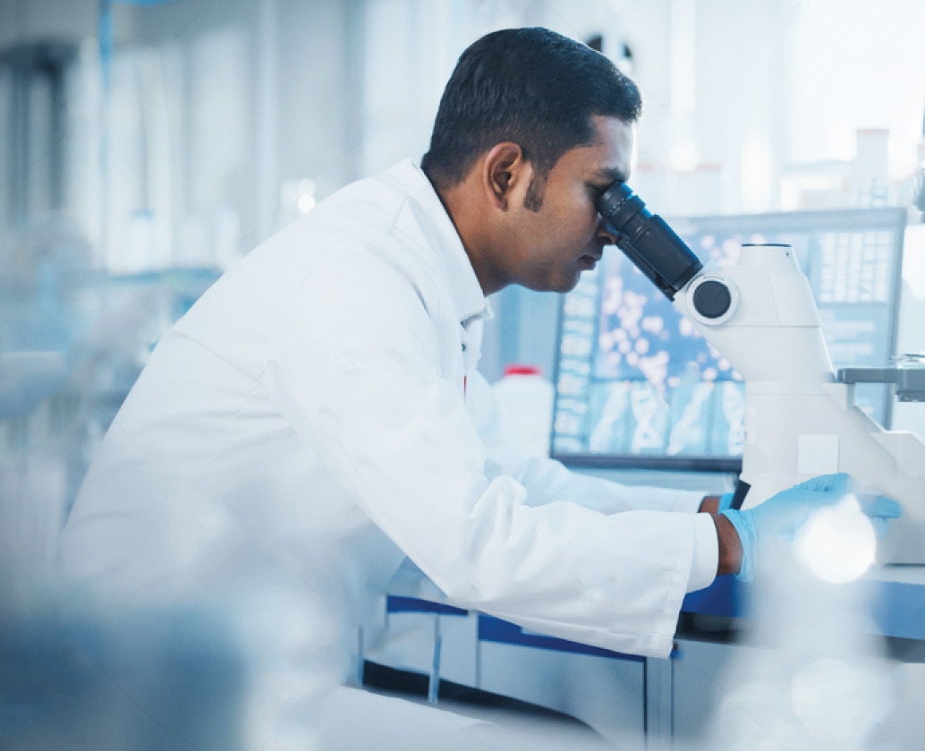 Male researcher in lab coat looking through a microscope beside computer monitors showing data