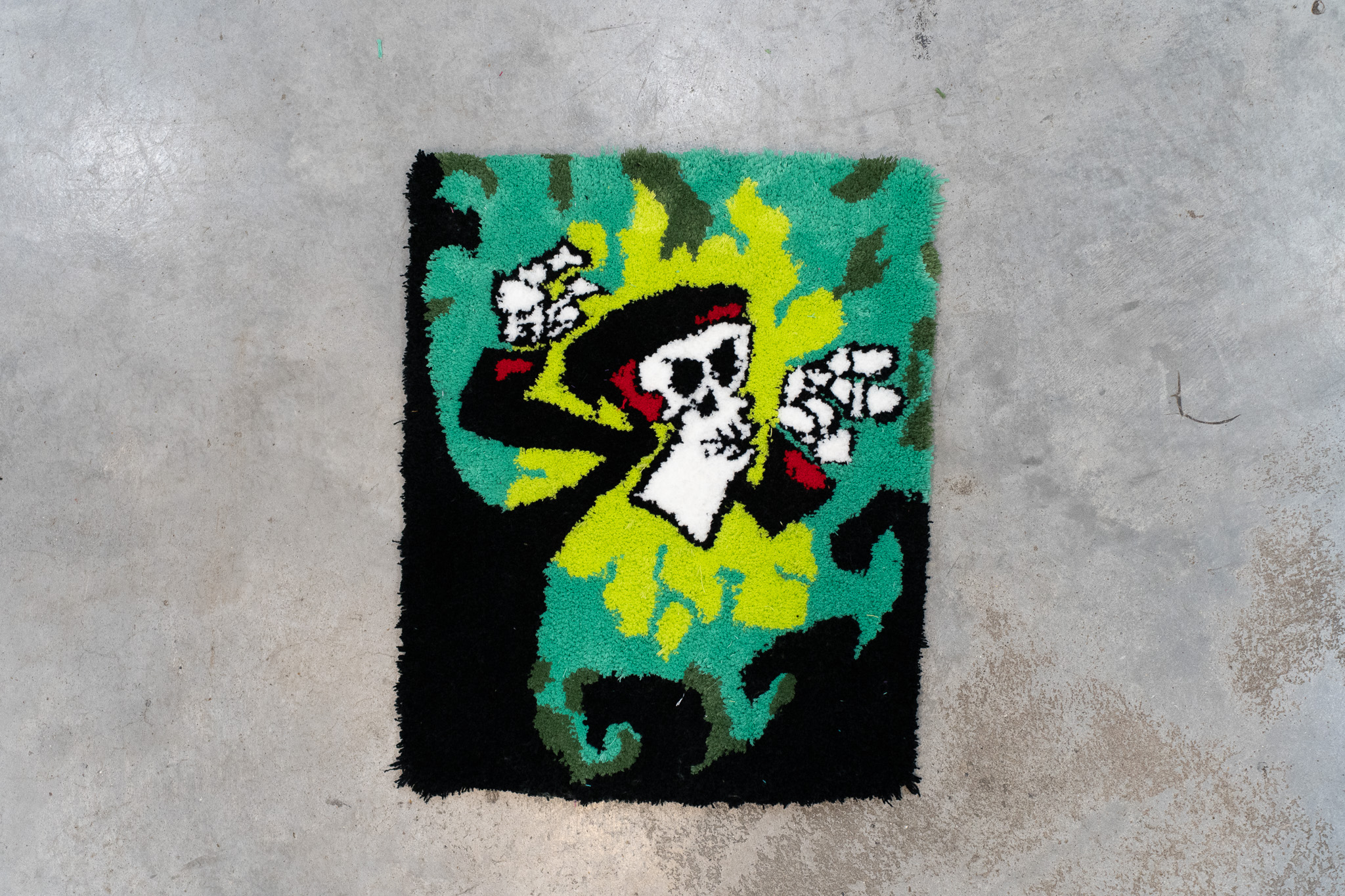 Colorful tufted textile artwork depicting a stylized skeleton figure with a black hat and hands raised, surrounded by green and yellow abstract patterns on a black background.