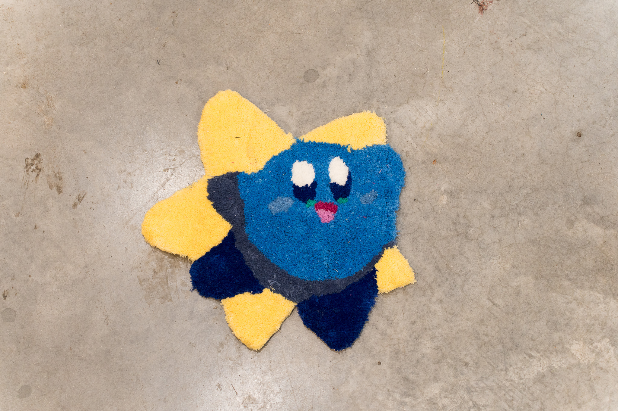 Colorful rug shaped like a smiling blue character with white eyes and a pink mouth on a yellow star background on a concrete floor.