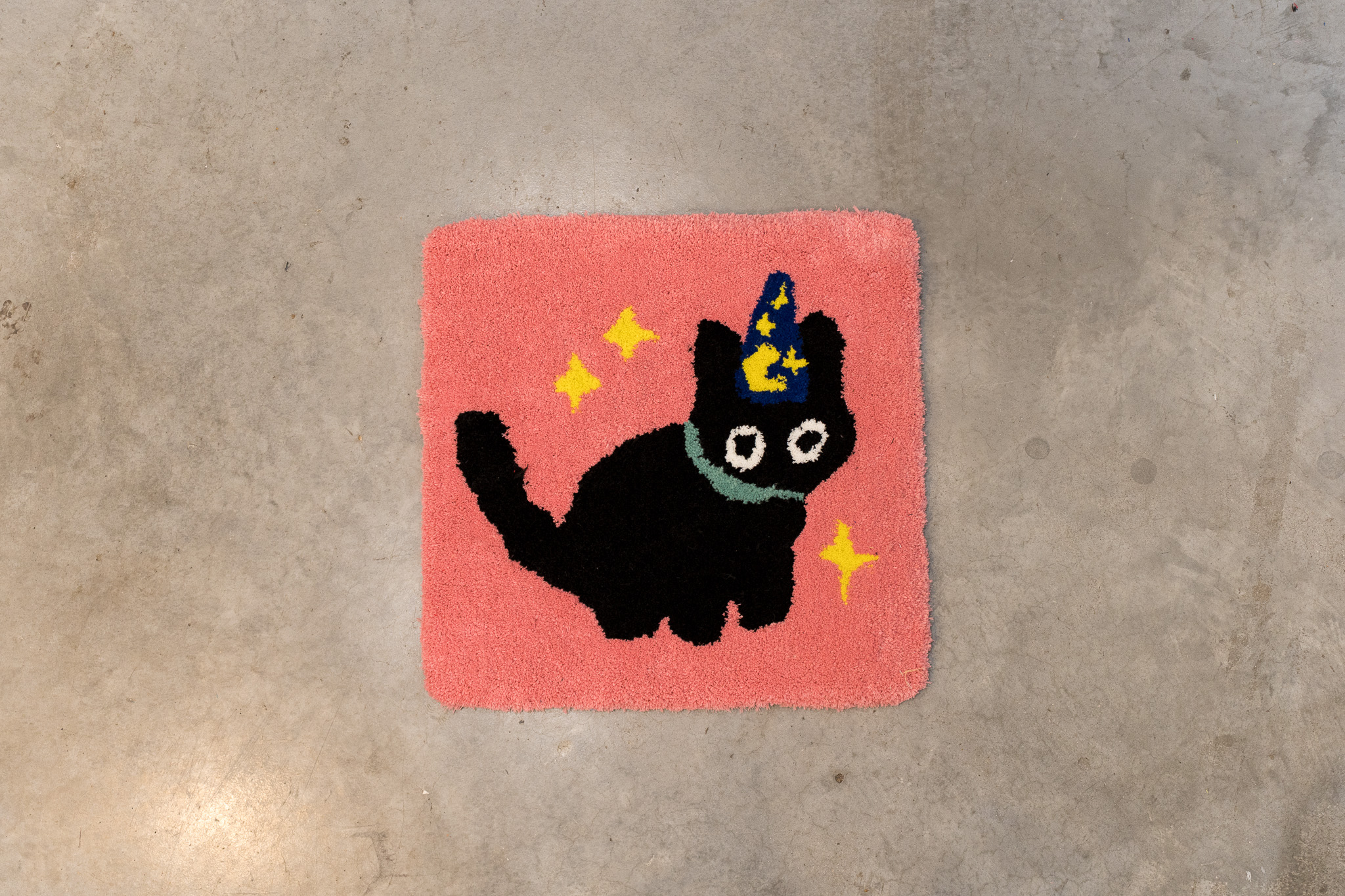 Square pink rug with a black cat wearing a blue and yellow wizard hat and a teal collar, surrounded by three yellow stars.
